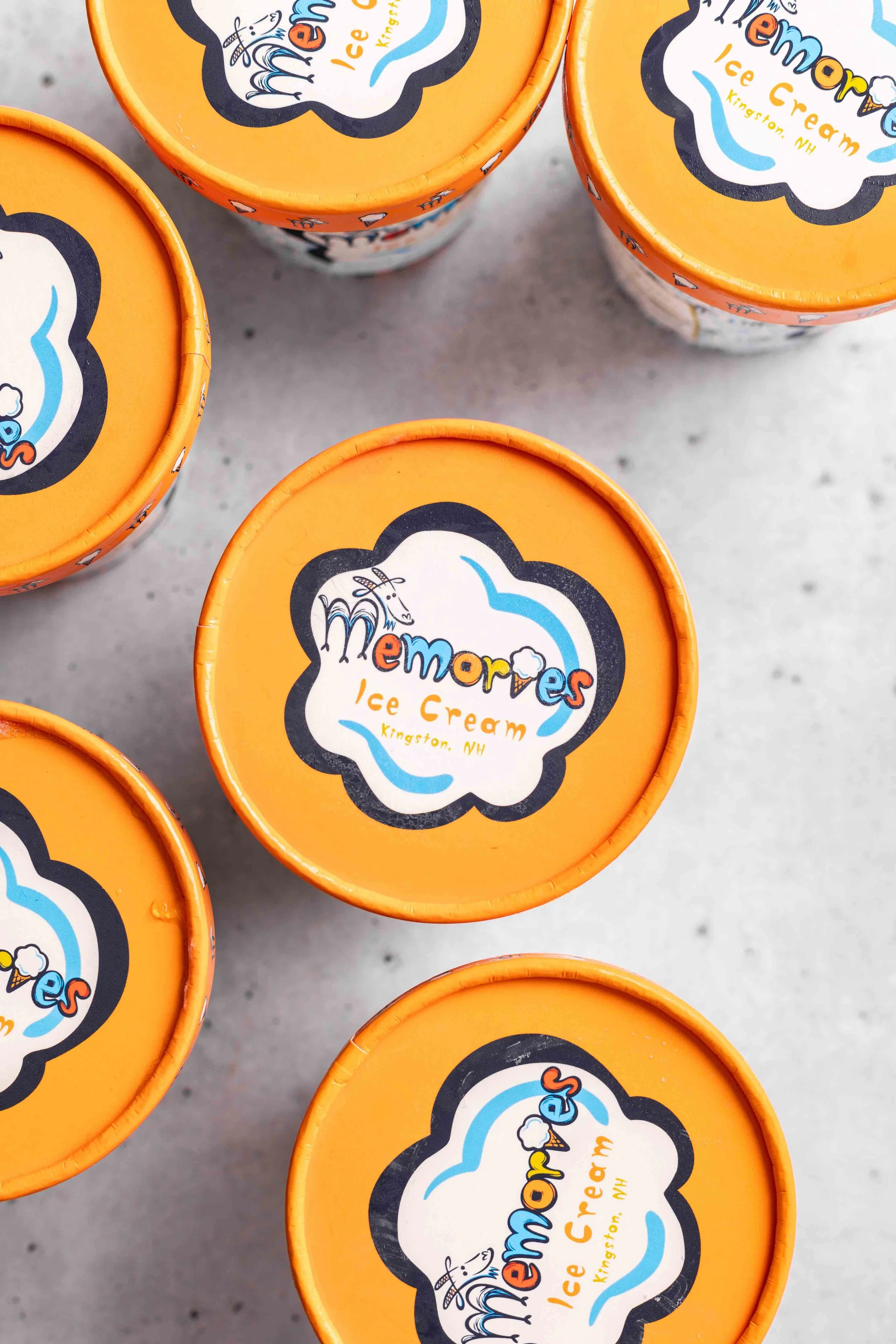 Frozen yogurt cups with orange lids, branded as Memories Ice Cream from Kingston, NH, featuring a cartoon ice cream cone and playful text on a gray surface.