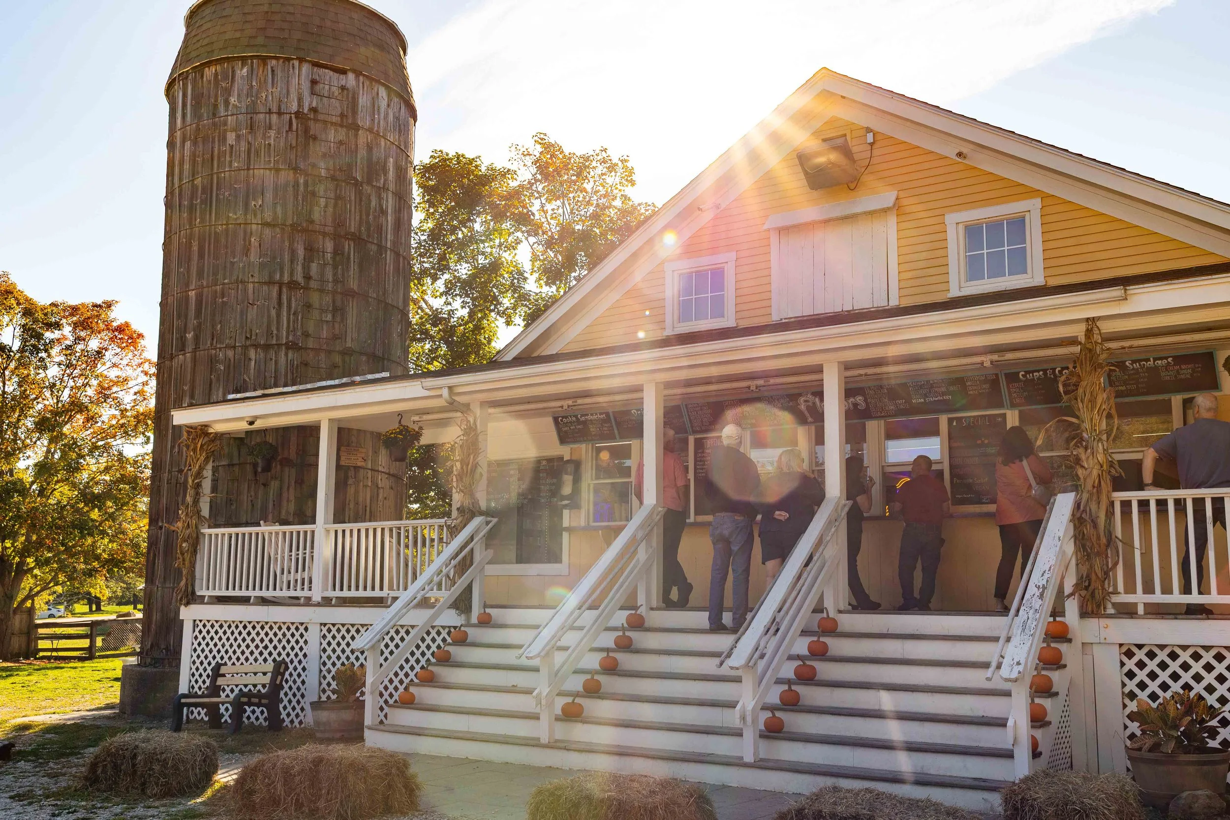 People line up at a counter on the porch of a yellow country store with a large wooden silo beside it, autumn trees in the background, and pumpkins decorating the stairs.