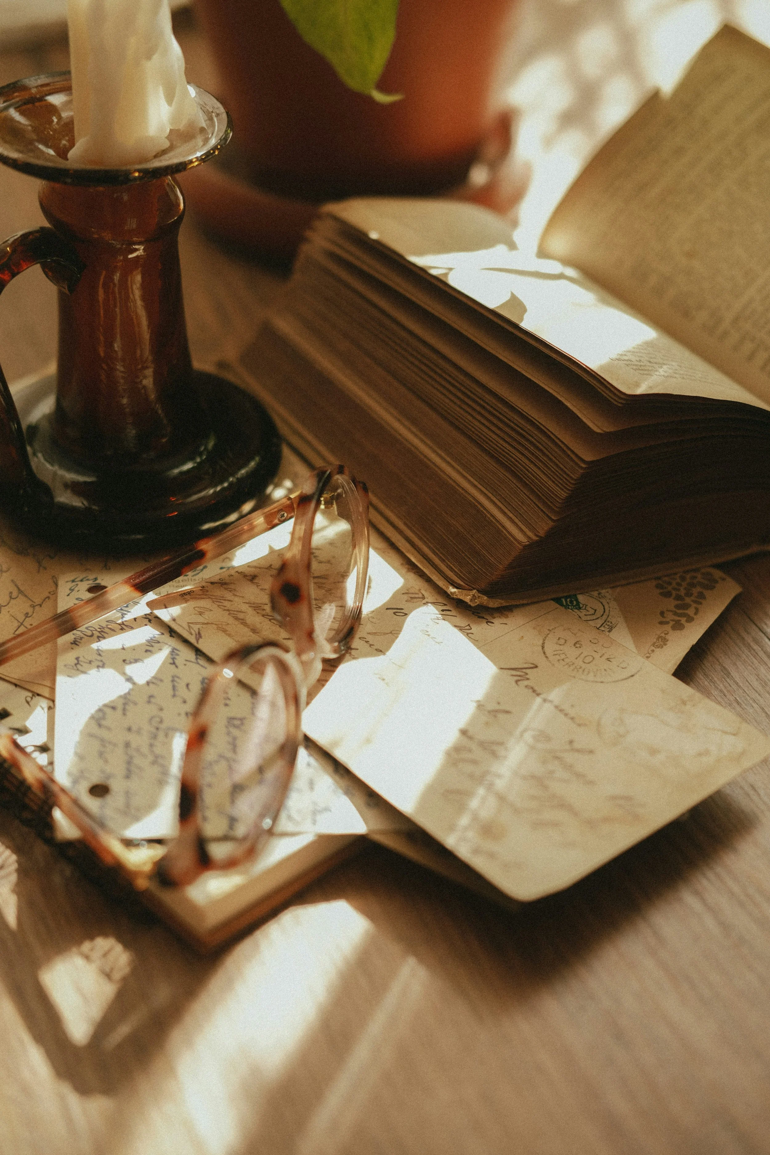 A wooden table holding an open book, worn photographs, a pair of tortoiseshell glasses, a small glass candle holder, and a potted plant.