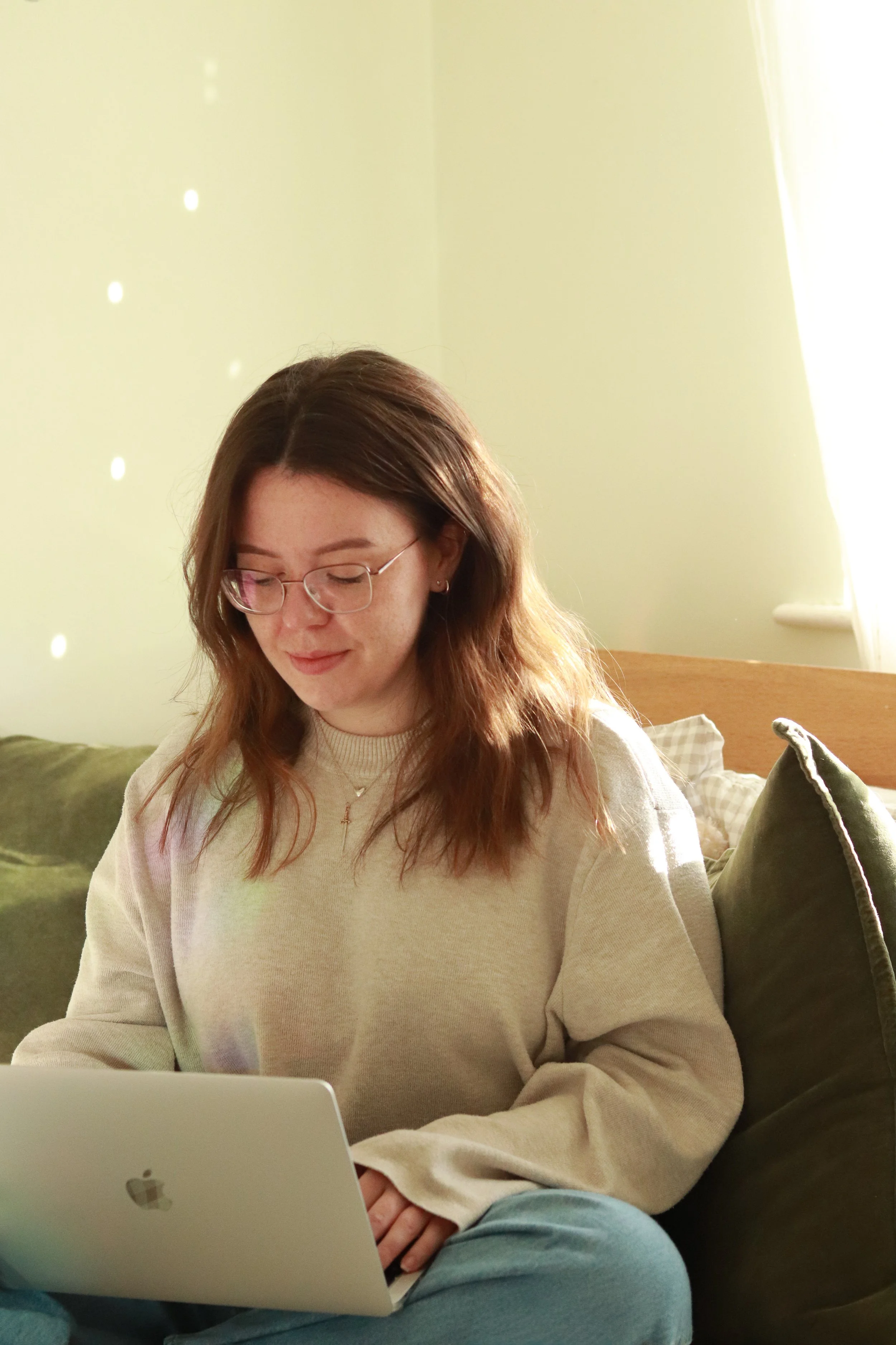 Young woman with glasses sitting on a green sofa, working on a MacBook in a sunlit room.