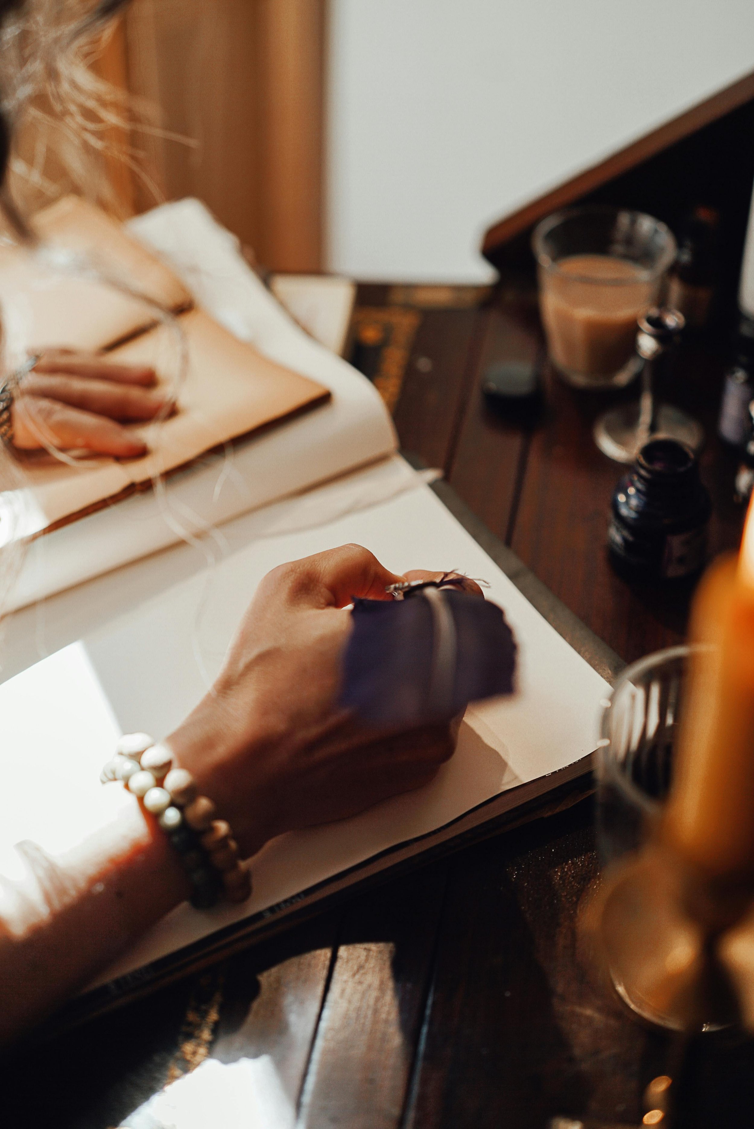 A person writing in a sketchbook at a wooden desk, surrounded by art supplies and a drink.