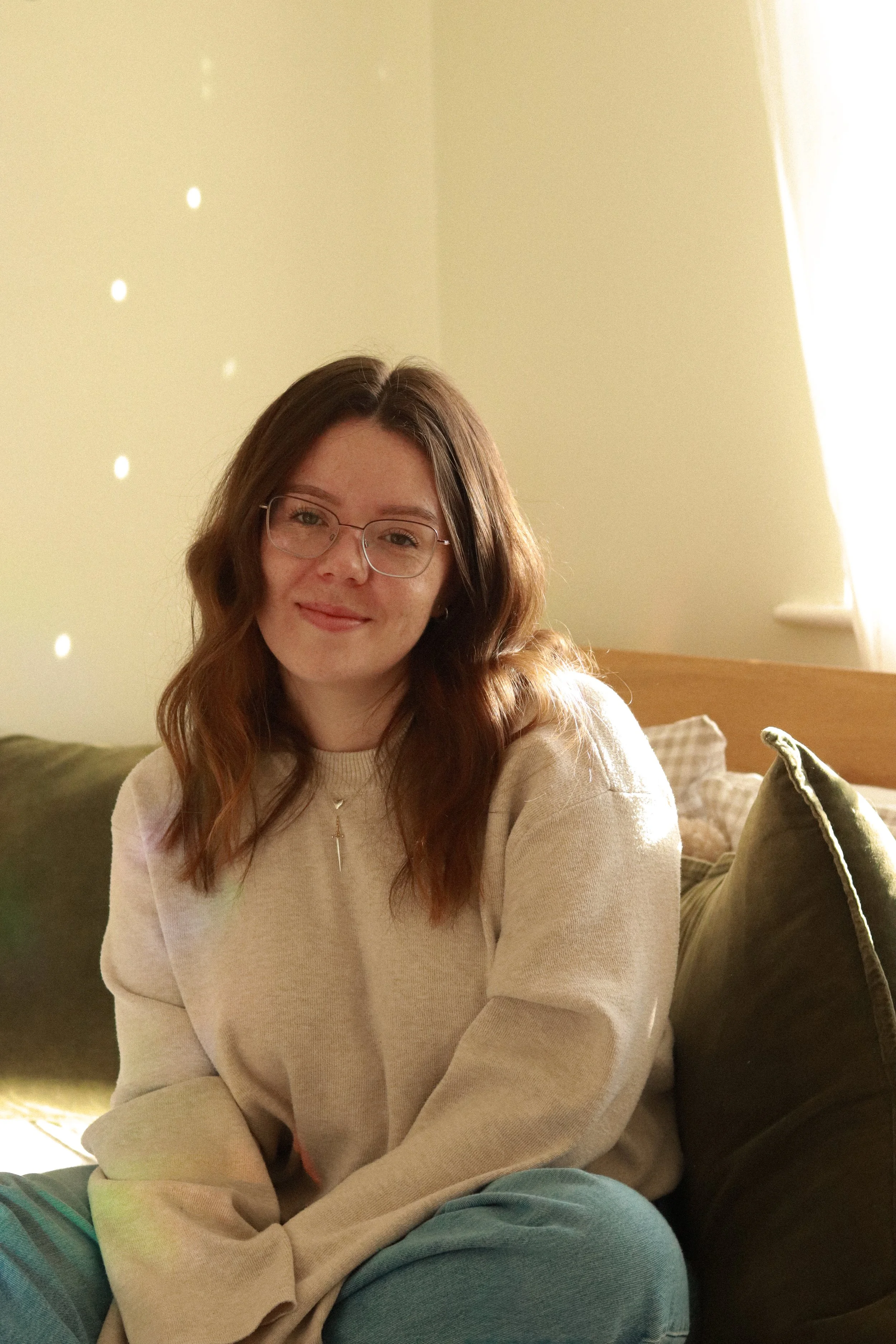 A young woman with long wavy brown hair and glasses sitting on a bed near a window, smiling softly in a well-lit room.
