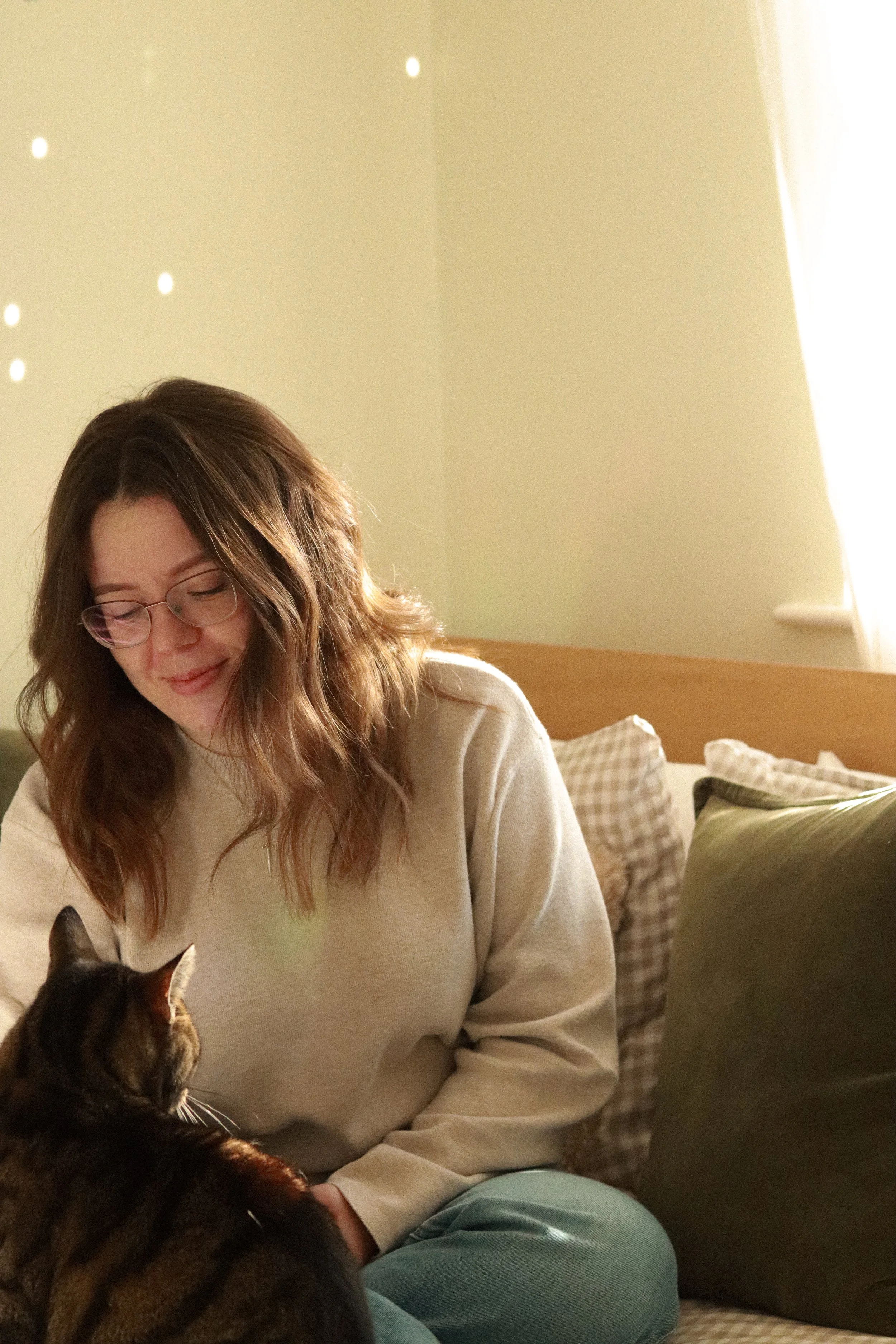 A woman with glasses and shoulder-length wavy brown hair sitting on a bed with a tabby cat in her lap, in a softly lit bedroom.