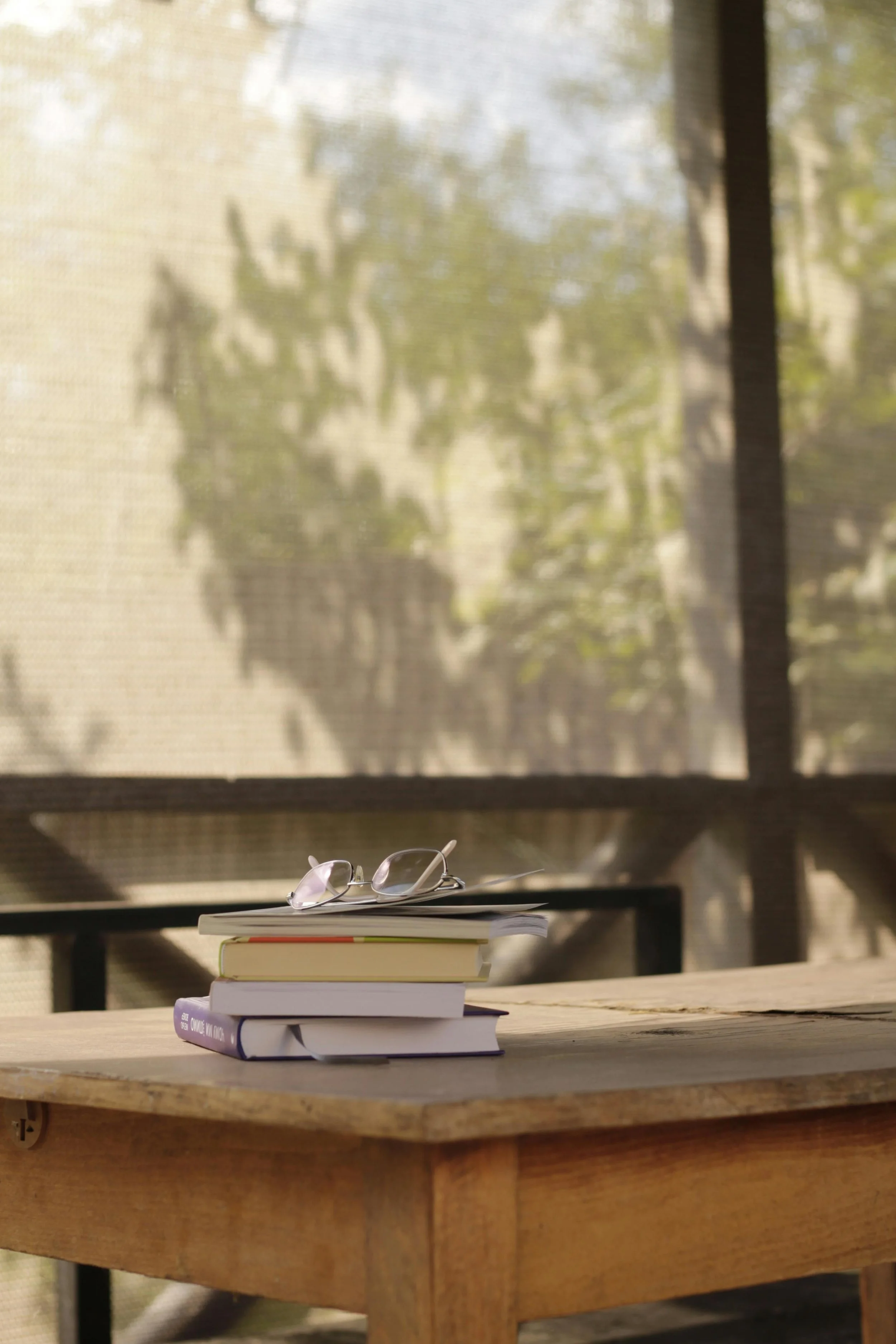 A stack of books with glasses on top sits on a wooden table near a window with sheer curtains, through which sunlight and blurry greenery are visible.
