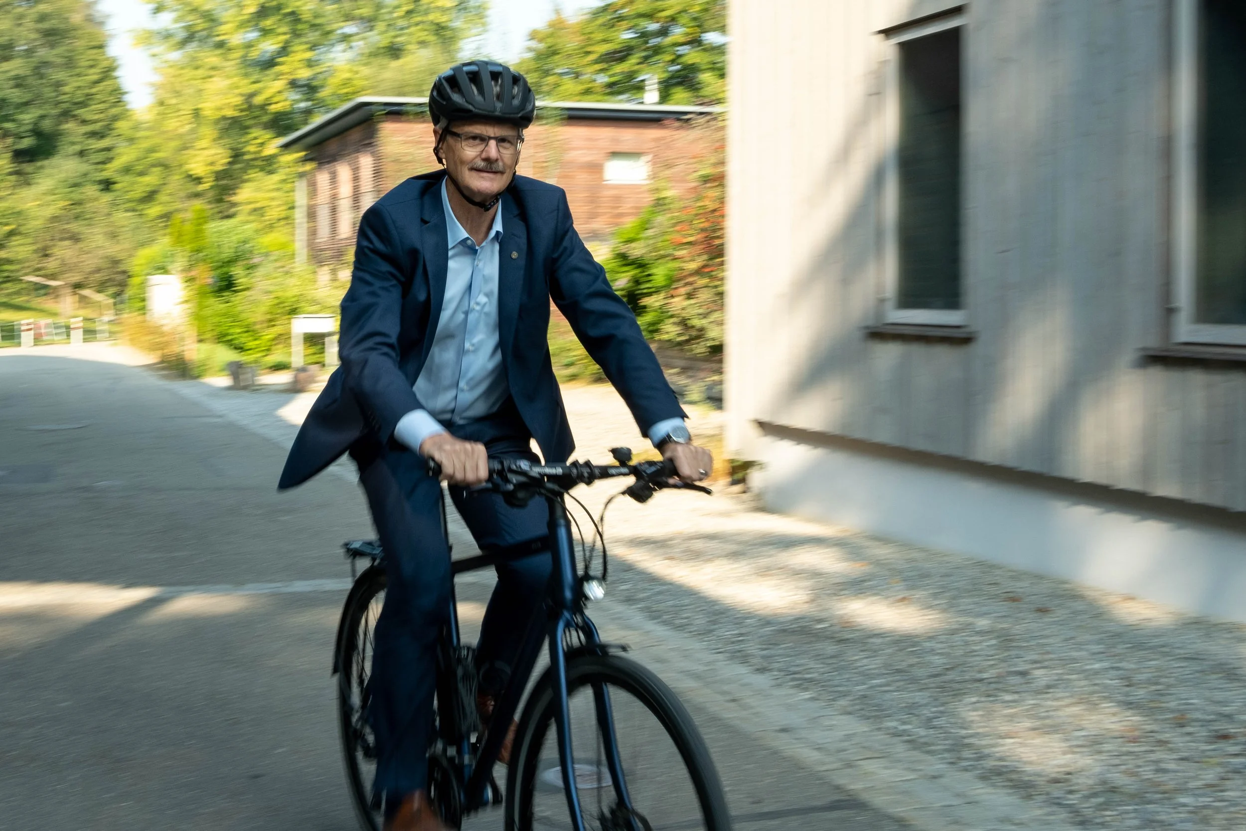 Ein Mann in Anzug und Fahrradhelm fährt auf einem Fahrrad durch eine Straße in einer ländlichen Gegend, umgeben von Bäumen und Gebäuden.