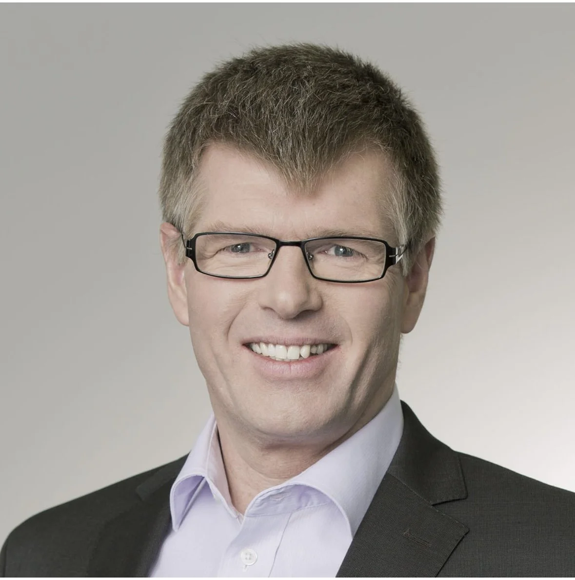 Porträt eines Mannes mit Brille, kurzem braunem Haar, weißem Hemd und schwarzer Jacke vor neutralem Hintergrund.