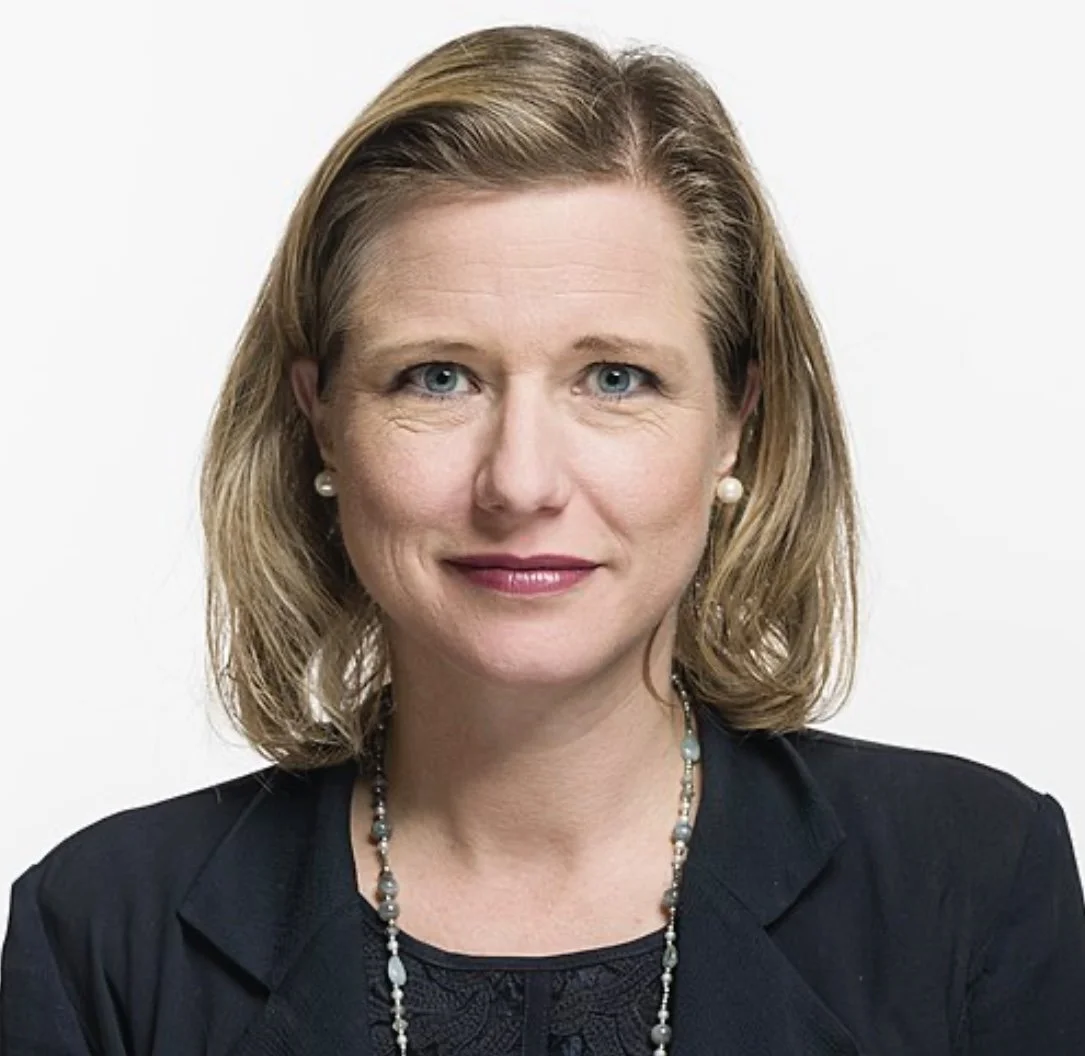 Porträt einer Frau mit blonden, lockigen Haaren, blauen Augen, trägt eine schwarze Jacke, Perlenohrringe und eine Perlenkette, vor weißem Hintergrund.