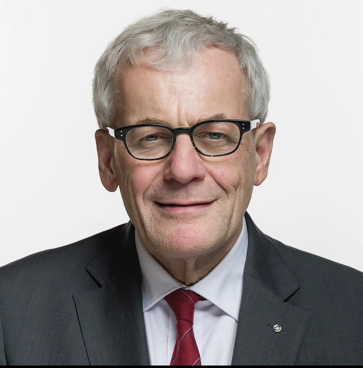 Porträt eines älteren Mannes mit Brille, Anzug, weißem Hemd und roter Krawatte, lächelnd vor weißem Hintergrund.