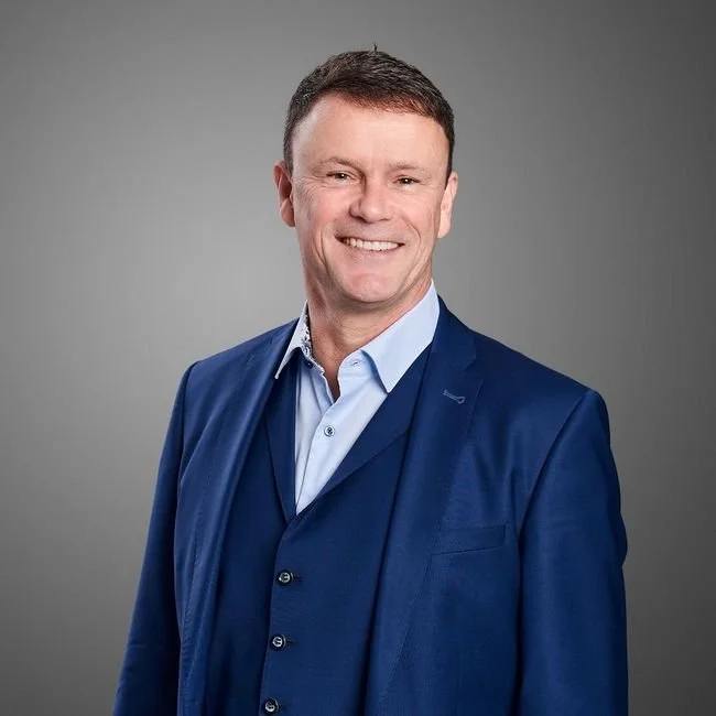 Ein Mann trägt einen blauen Anzug und ein hellblaues Hemd, lächelt vor grauem Hintergrund.