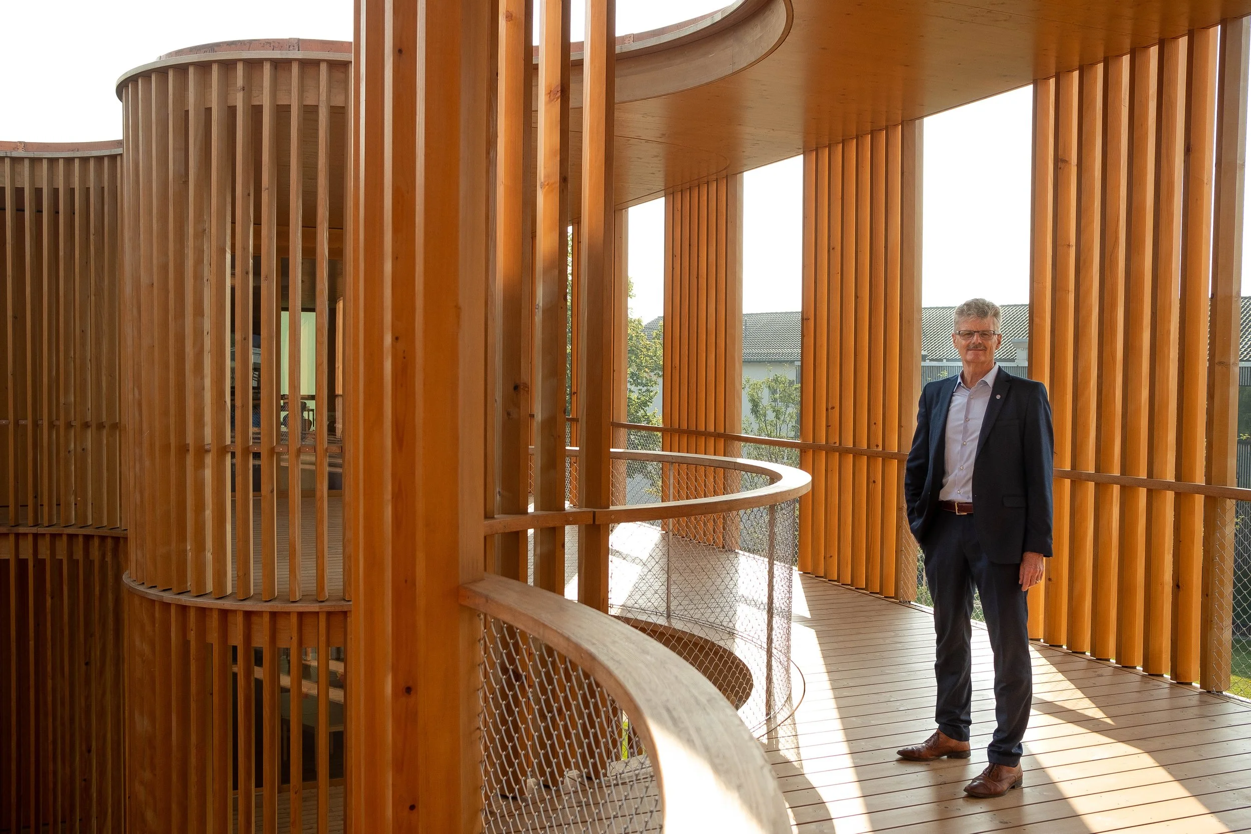 Ein Mann in formeller Kleidung steht auf einer Holzterrasse mit schmalen Holzbalustraden, umgeben von vertikalen Holzbrettern, mit einem Gebäude im Hintergrund.