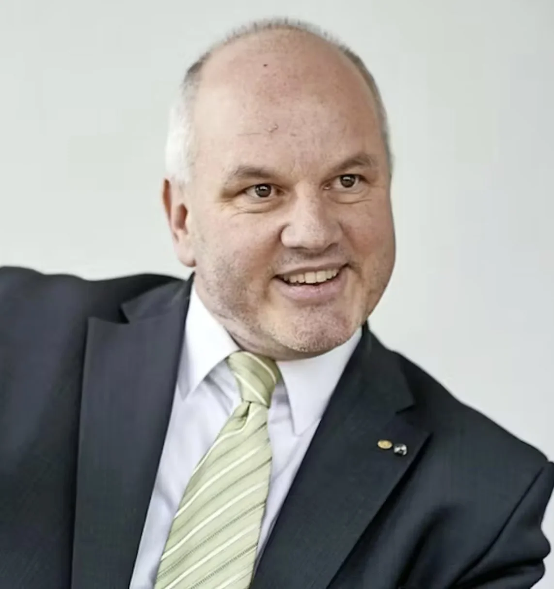 Ein lachender Mann in formeller Kleidung, schwarzer Anzug, weißes Hemd und grüner Krawatte, sitzt in einem Büro.