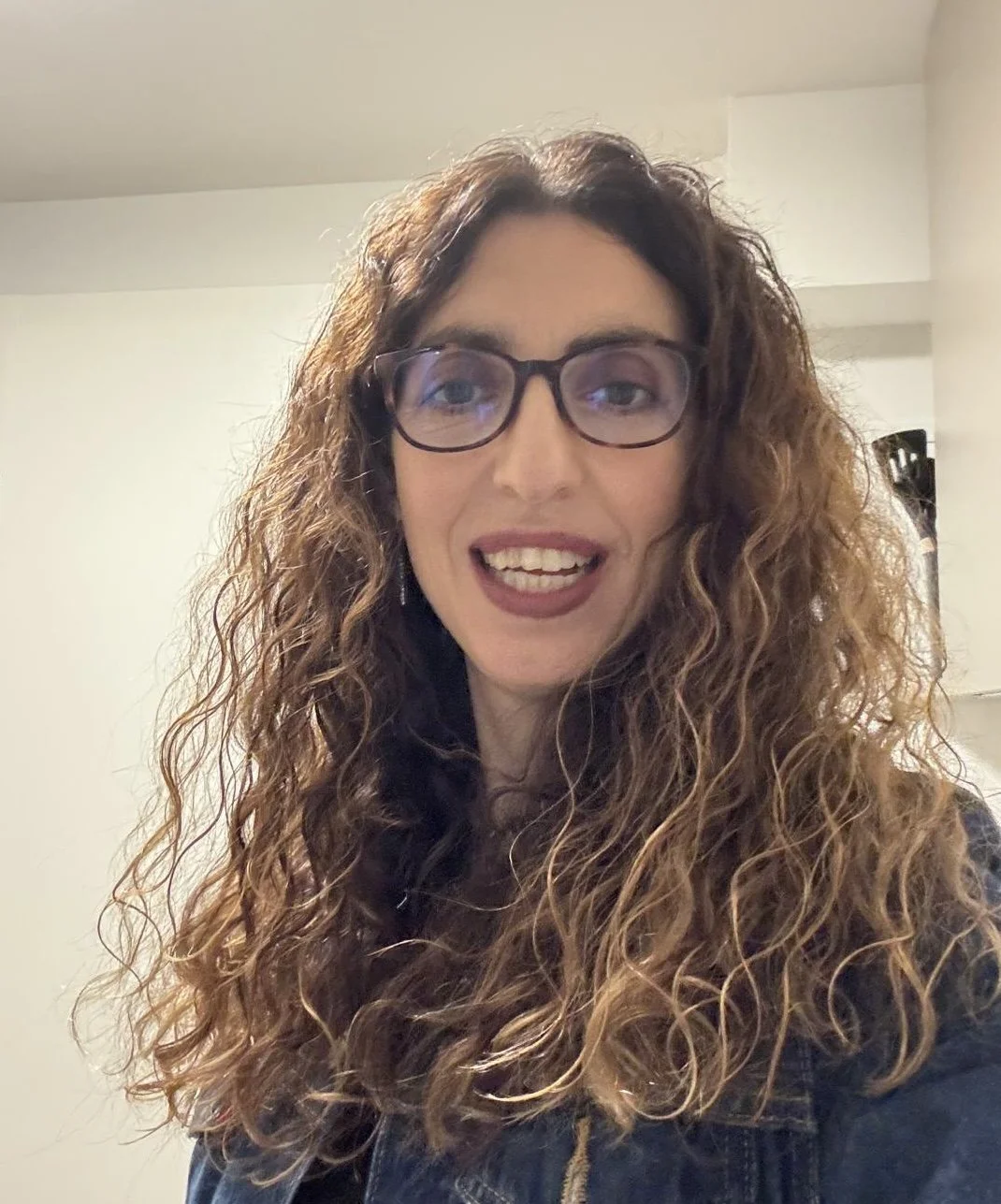 Eine Frau mit lockigem braunem Haar, Brille und dunklem Lippenstift lächelnd vor einem neutralen Hintergrund.