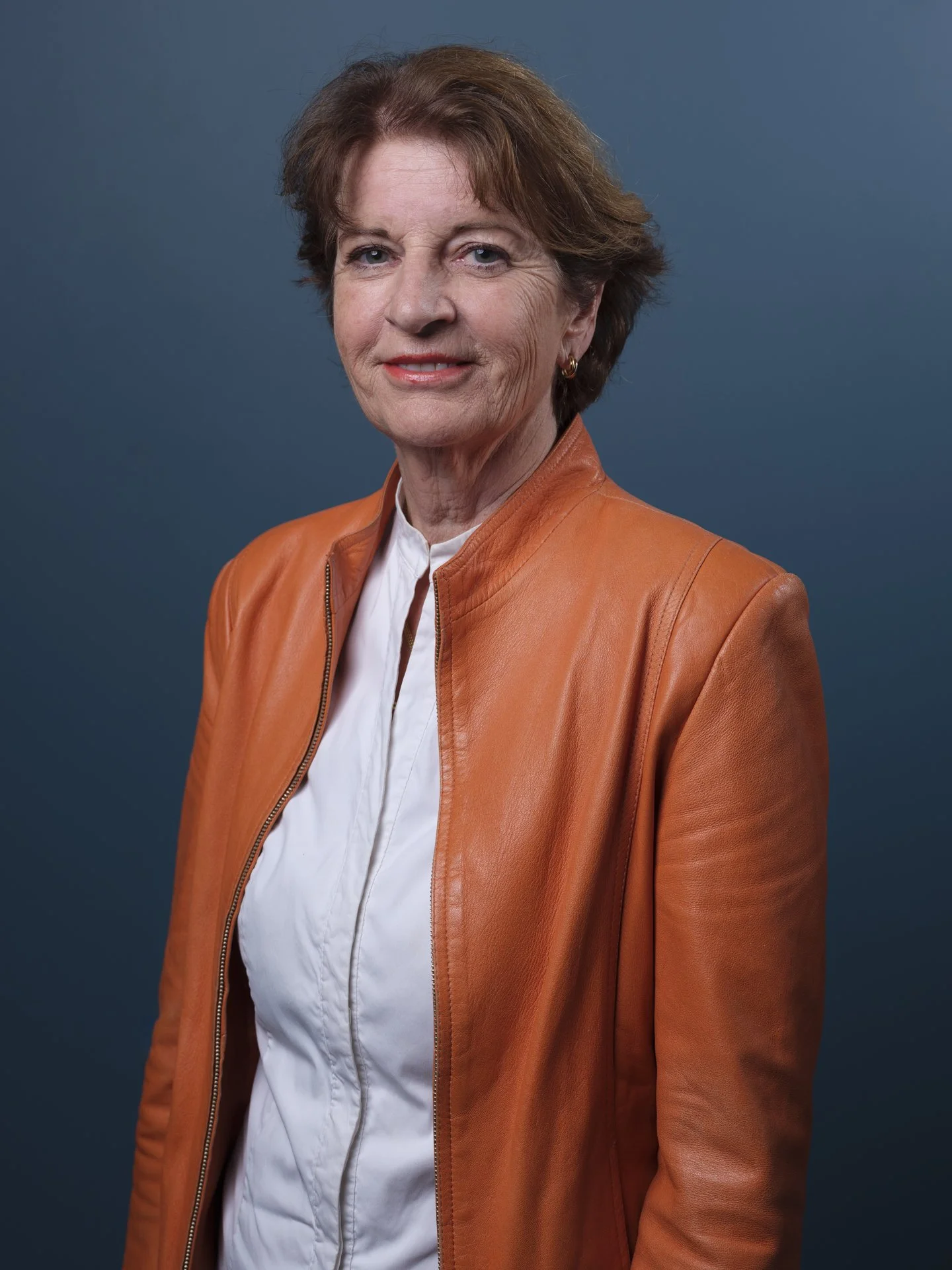Porträt einer älteren Frau mit kurzen braunen Haaren, trägt eine orange Lederjacke und ein weißes Hemd, vor dunkelblauem Hintergrund.