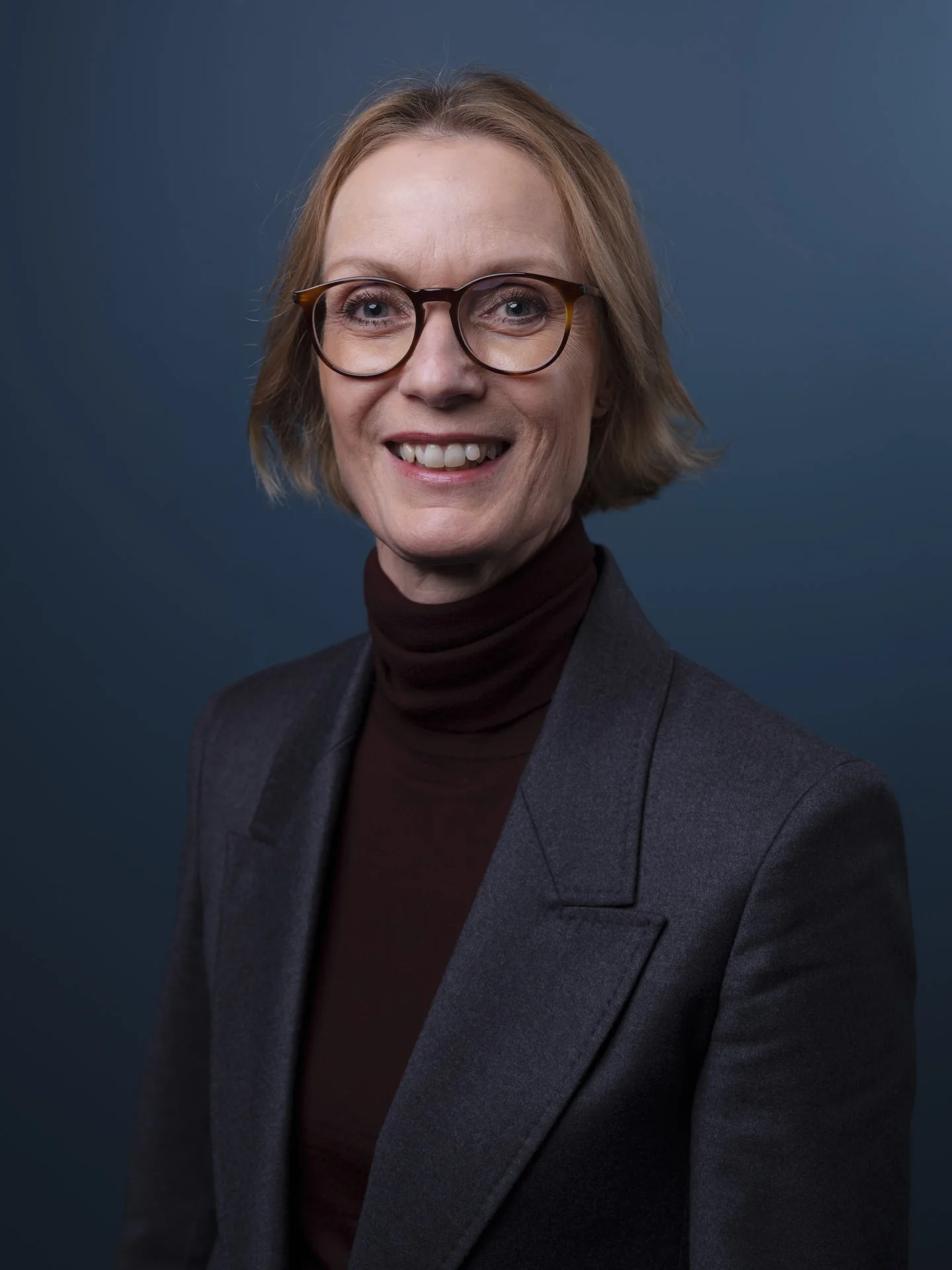 Porträt einer lächelnden Frau mit Brille, braunem Turtleneck und dunklem Blazer vor blauem Hintergrund.