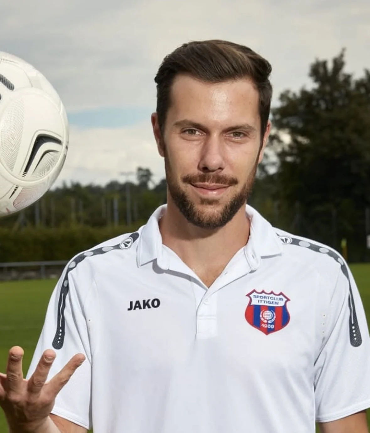 Ein junger Mann in einem weißen Sporttrikot, der auf einem Fußballfeld steht, während er einen Fußball in der Nähe der Kamera hält.