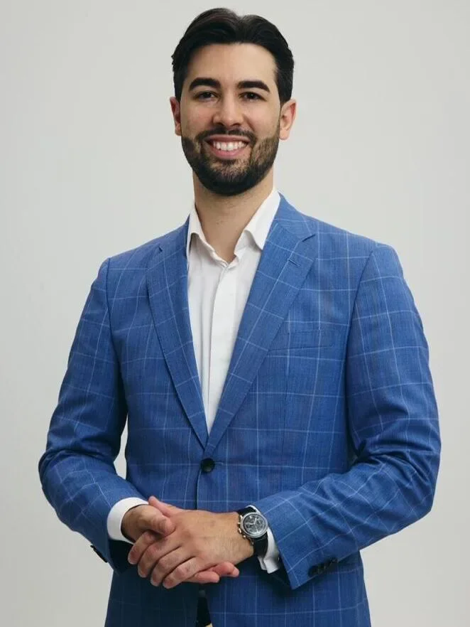 Ein Mann trägt einen blauen Anzug mit kariertem Muster und ein weißes Hemd, hat dunkle Haare und trägt einen Bart. Er lächelt und steht vor einem neutralen Hintergrund.