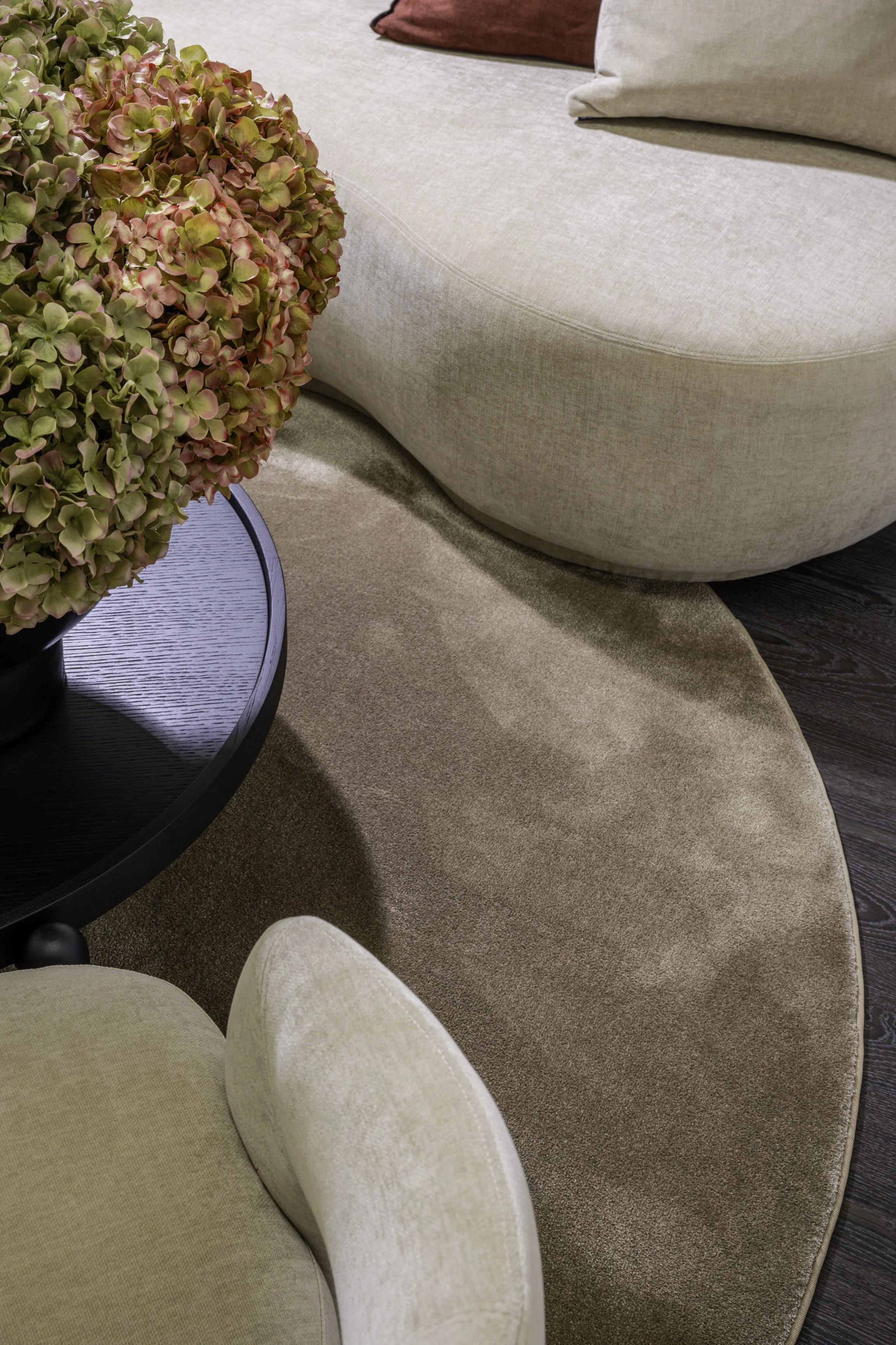 Nahaufnahme von einem Wohnzimmer mit Blumen auf einem schwarzen Tisch, beige Sessel, Couch und einem runden Teppich.