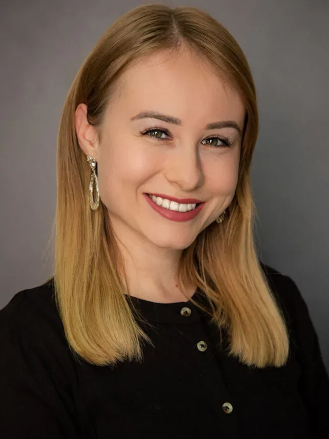 Lächelnde junge Frau mit blonden Haaren, roten Lippen, Ohrringen und schwarzem Oberteil vor grauem Hintergrund.