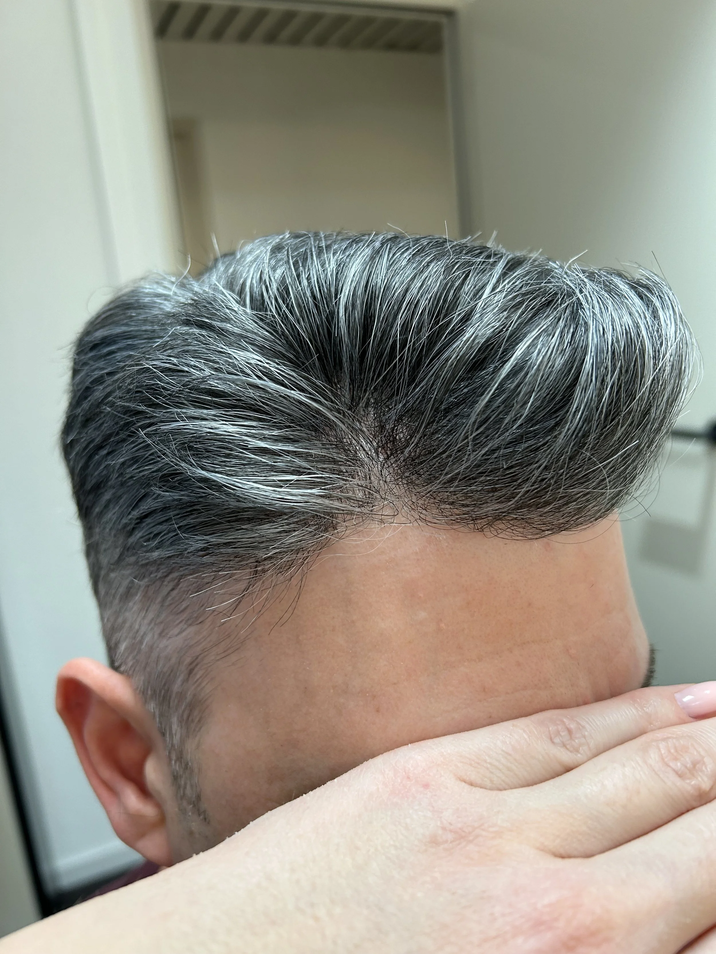 Nahaufnahme eines Mannes mit grauem, gepflegtem Haar, das nach oben gestylt ist, vor einem Spiegel in einem Badezimmer.