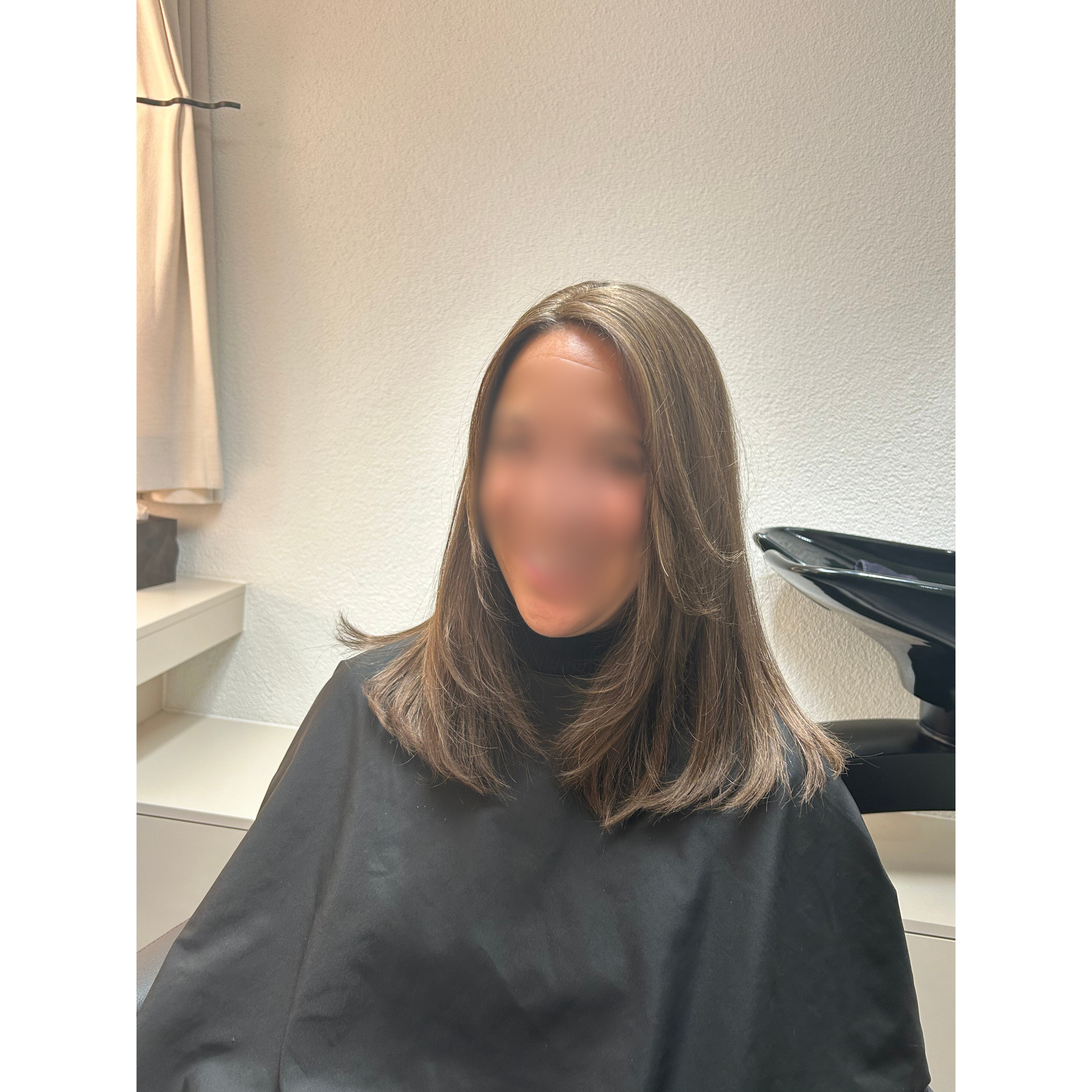 Eine Frau mit braunen, mittellangen Haaren trägt einen schwarzen Umhang, sitzt im Friseursalon, hinter ihr eine weiße Wand und ein Frisiertisch mit Strähnen.