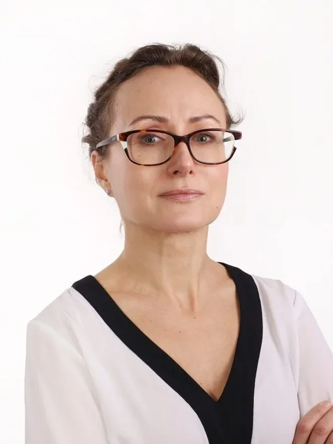 Eine Frau mit Brille, die einen weißen Oberteil mit schwarzem Rand trägt, schaut ernst in die Kamera.