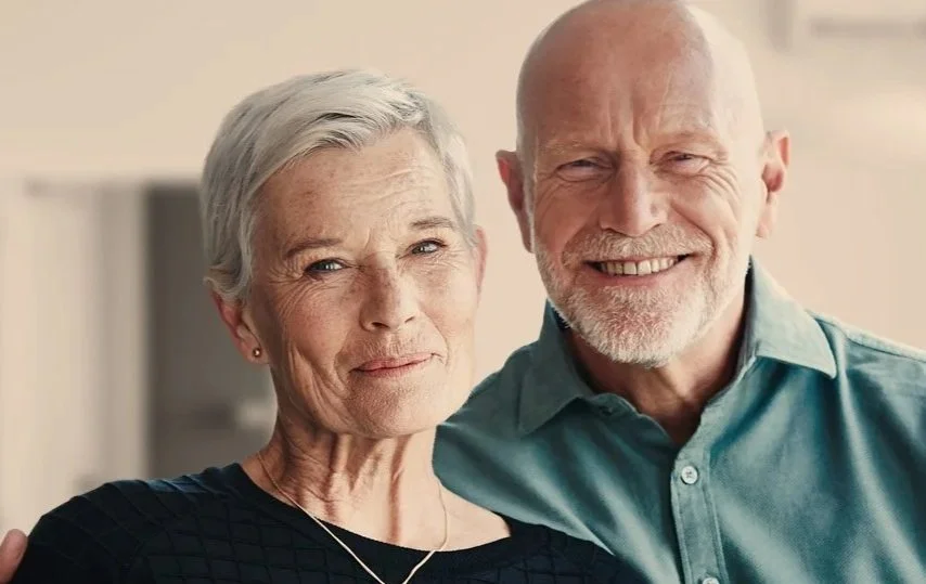 Ein älteres Paar, eine Frau mit kurzen grauen Haaren und ein Mann mit Glatze und grauem Bart, lachen und stehen dicht beieinander in einem hellen Raum.