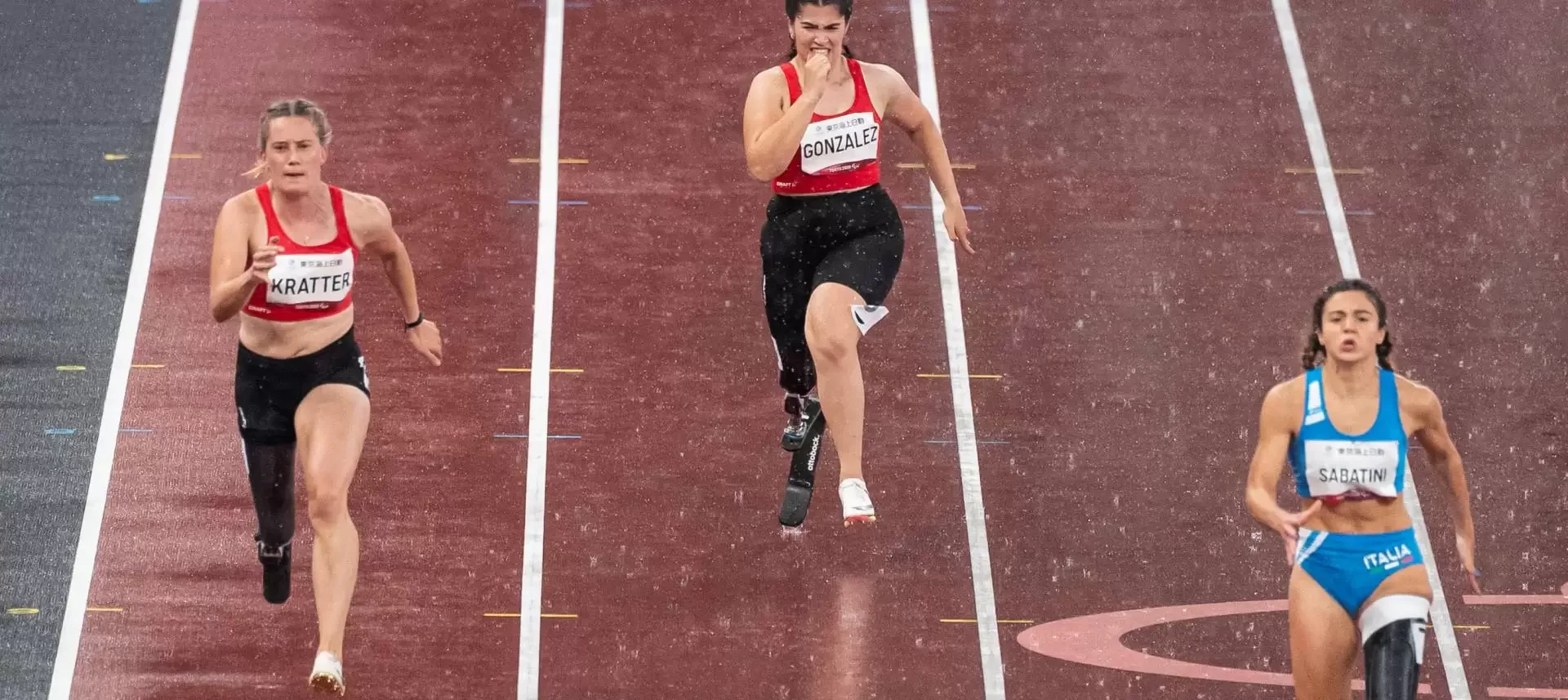 Drei weibliche Sprinterinnen beim Start eines Kurzstreckenlaufs auf nassem Stadionbelag, während es regnet.