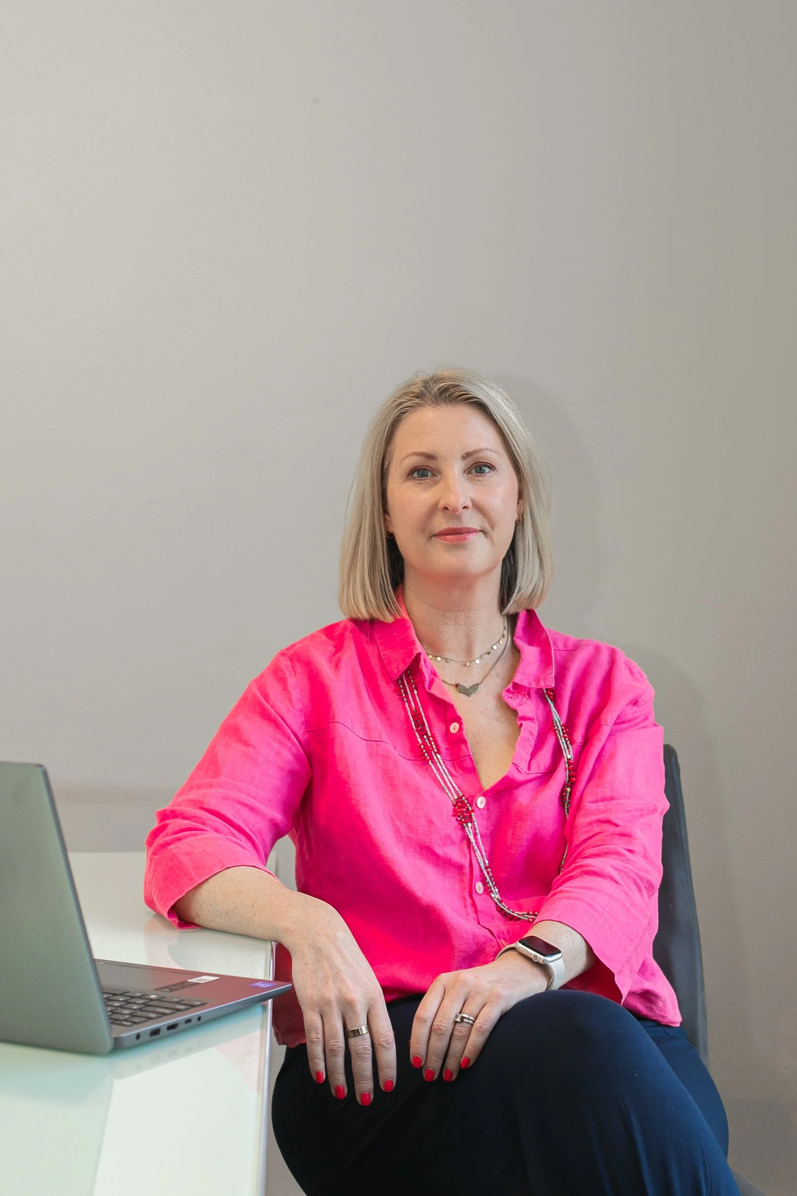 Woman with blonde hair wearing a pink blouse and black pants sitting at a table with a laptop in front of her, against a plain wall background.