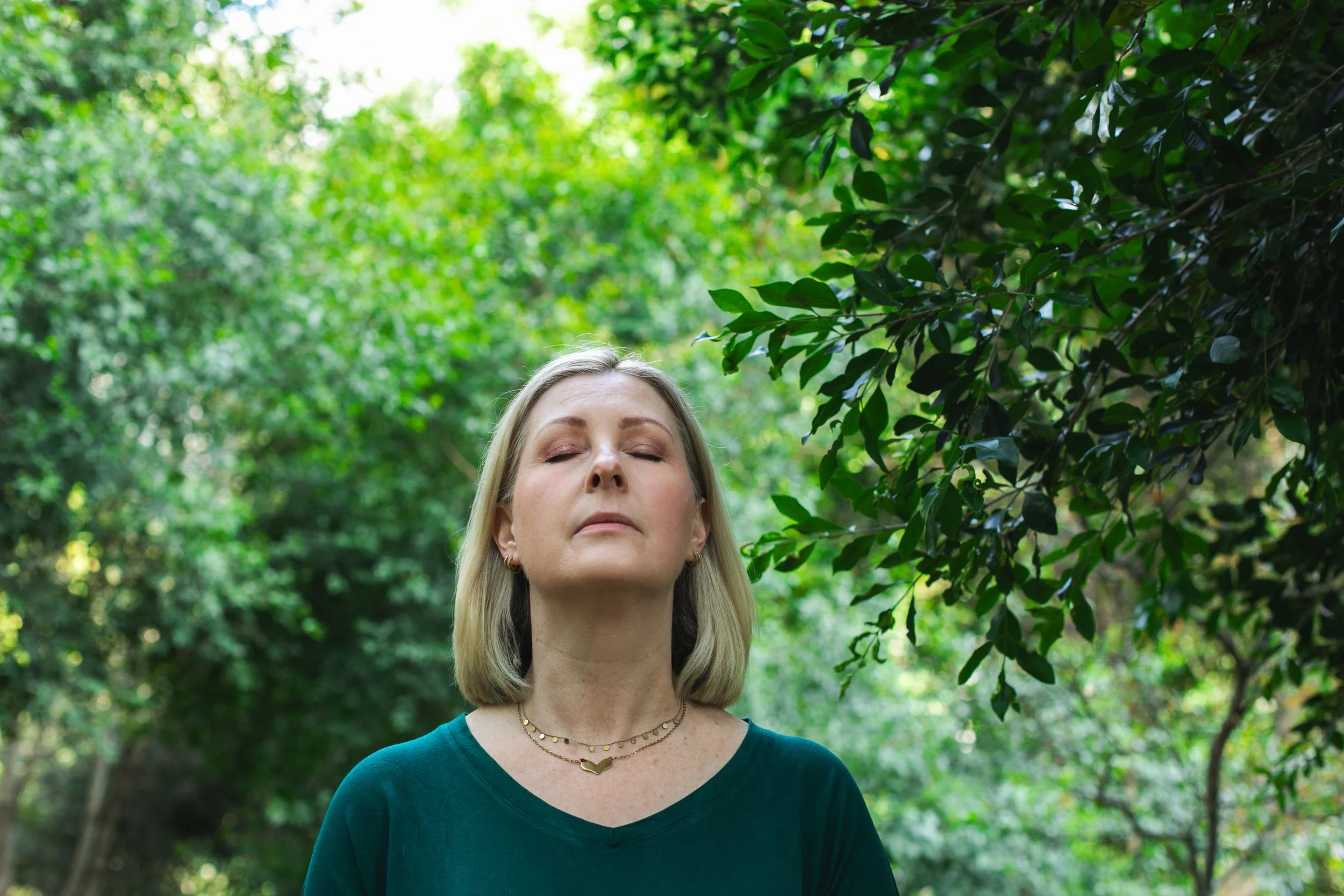 A woman with blonde hair and closed eyes standing outdoors in a lush green forest, wearing a dark green shirt and layered gold necklaces.