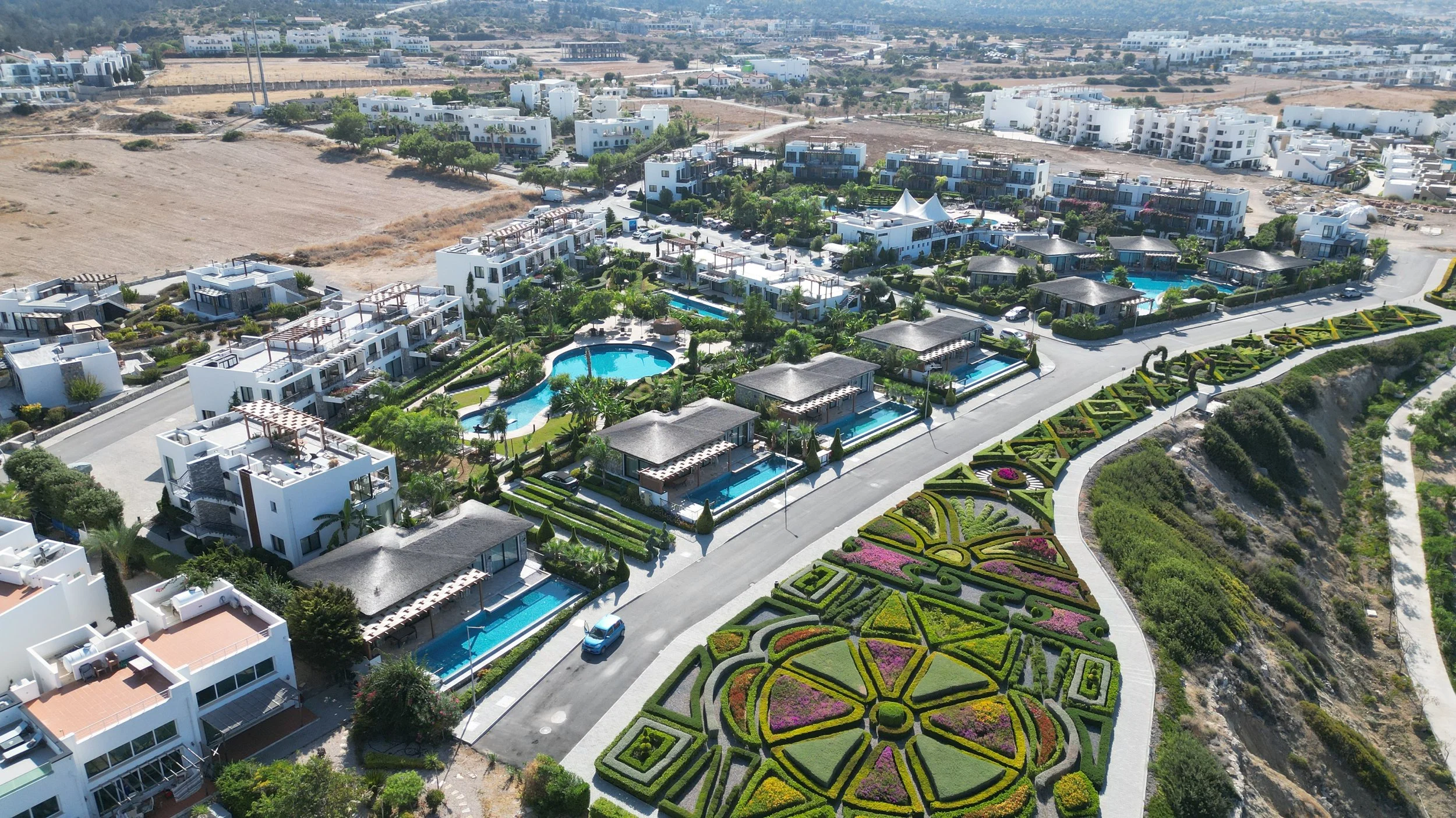An aerial view of a luxurious residential complex with white buildings, swimming pools, landscaped gardens, and a decorative flower garden, set against a semi-arid landscape.