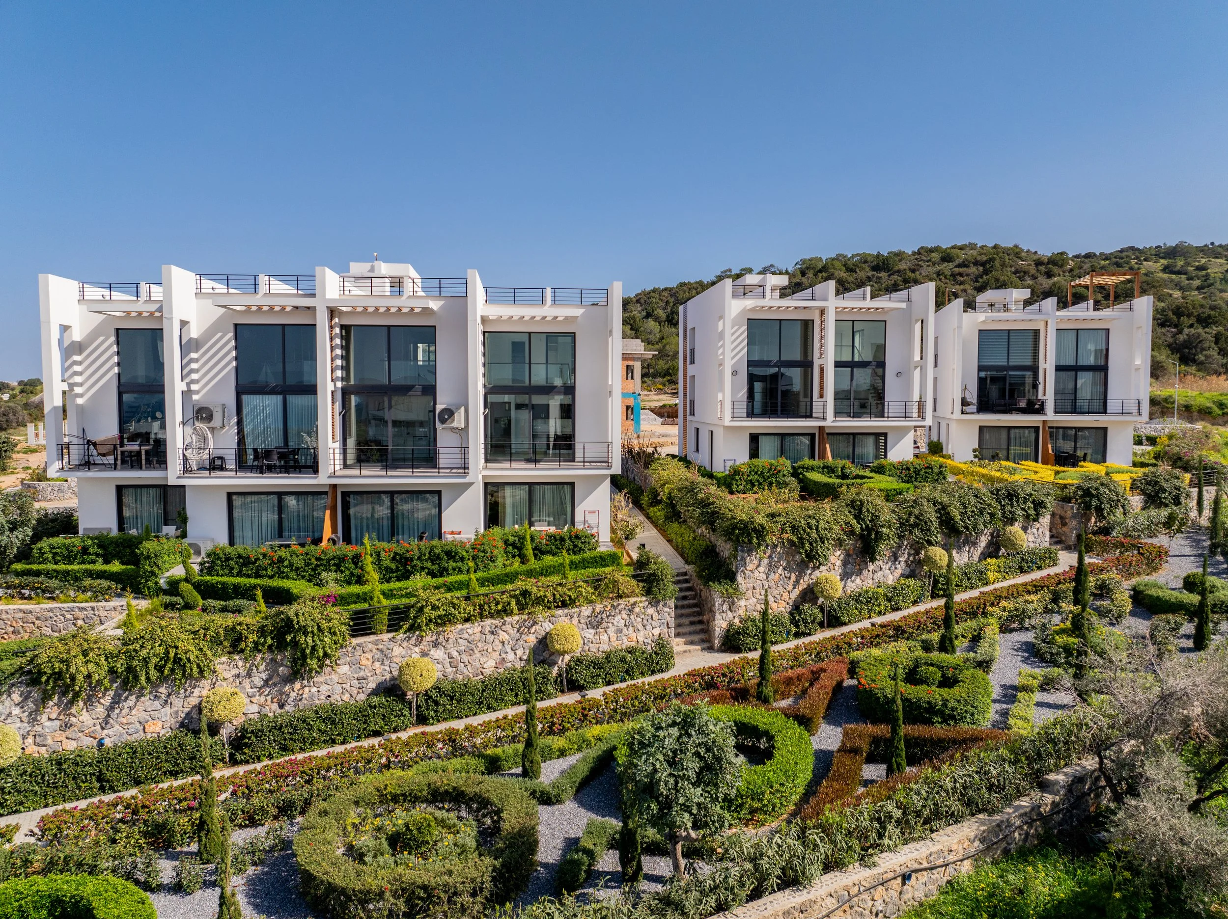 Modern white multi-story residential buildings with large glass windows and balconies, surrounded by landscaped gardens and terraced stone pathways under a clear blue sky.