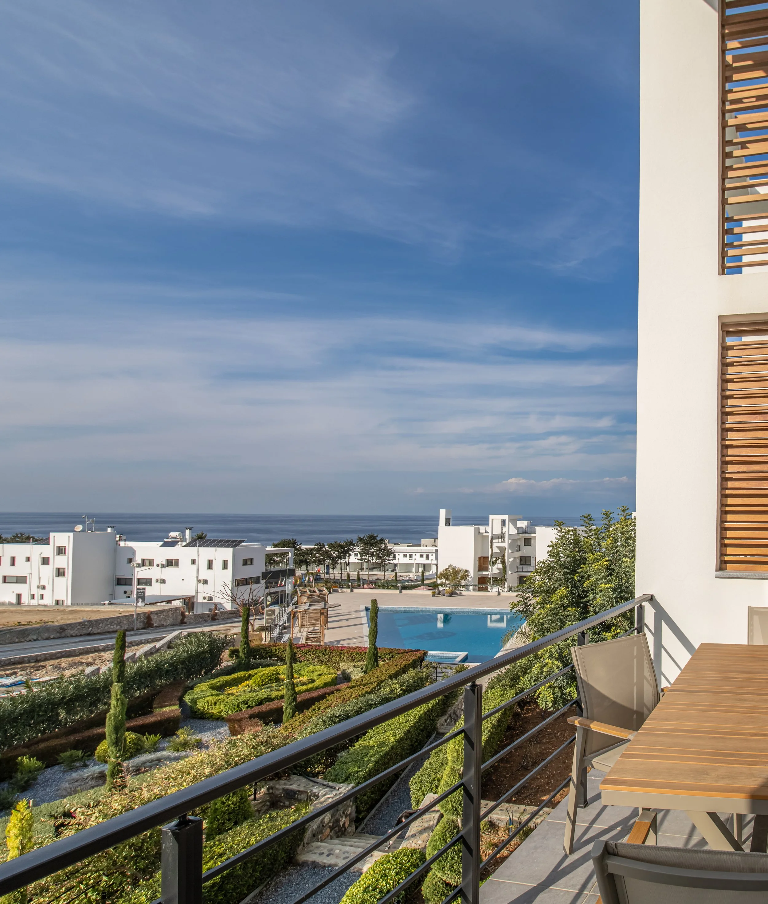 Balcony overlooking a swimming pool and landscaped garden with white buildings and ocean in the distance under a blue sky