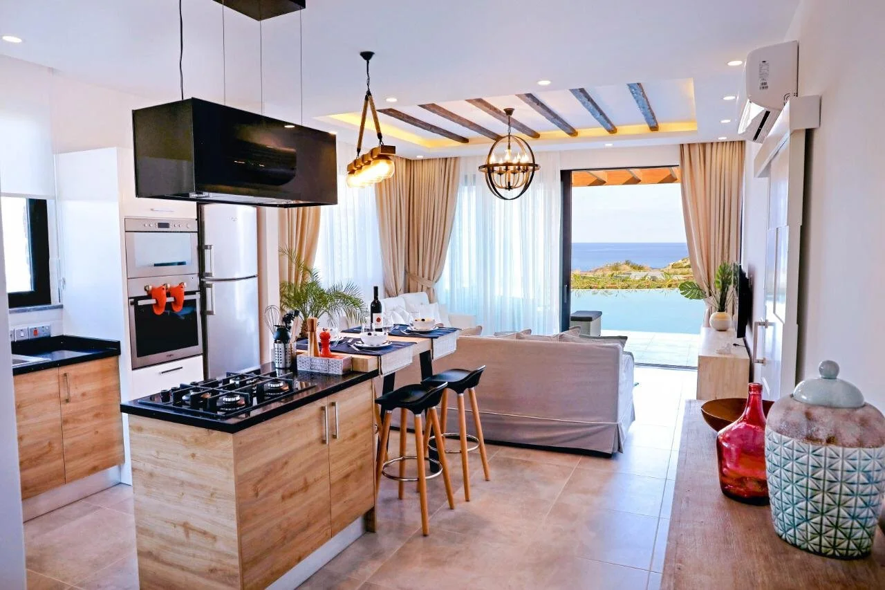 Open-concept living room and kitchen with ocean view, featuring a dining area, white sofas, potted plants, decorative vases, a mounted television, and sliding glass doors leading to a balcony.