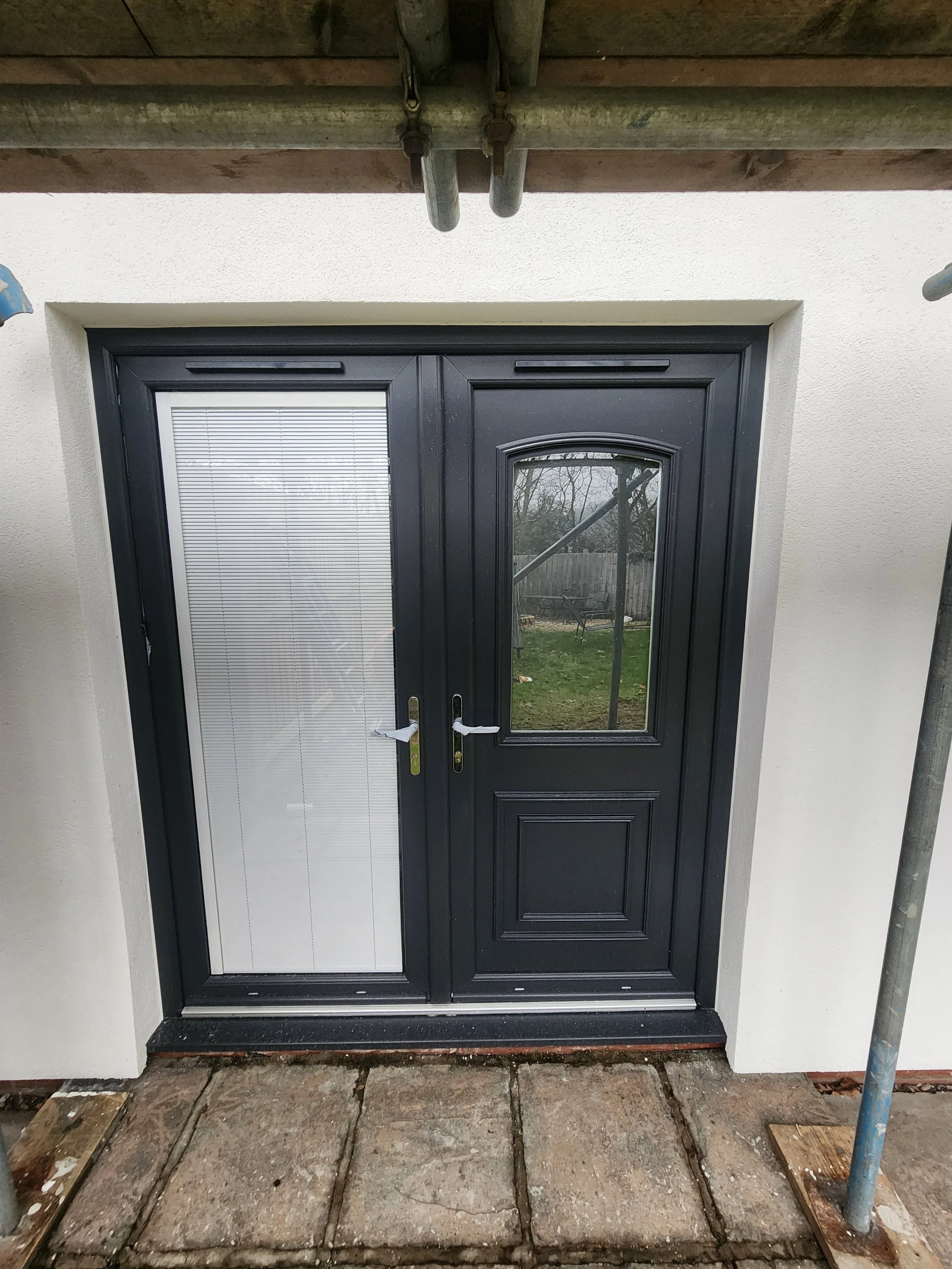 Exterior view of a black double door with a glass window pane on the right side. The door is set in a white wall, with construction scaffolding visible at the top and on the sides.