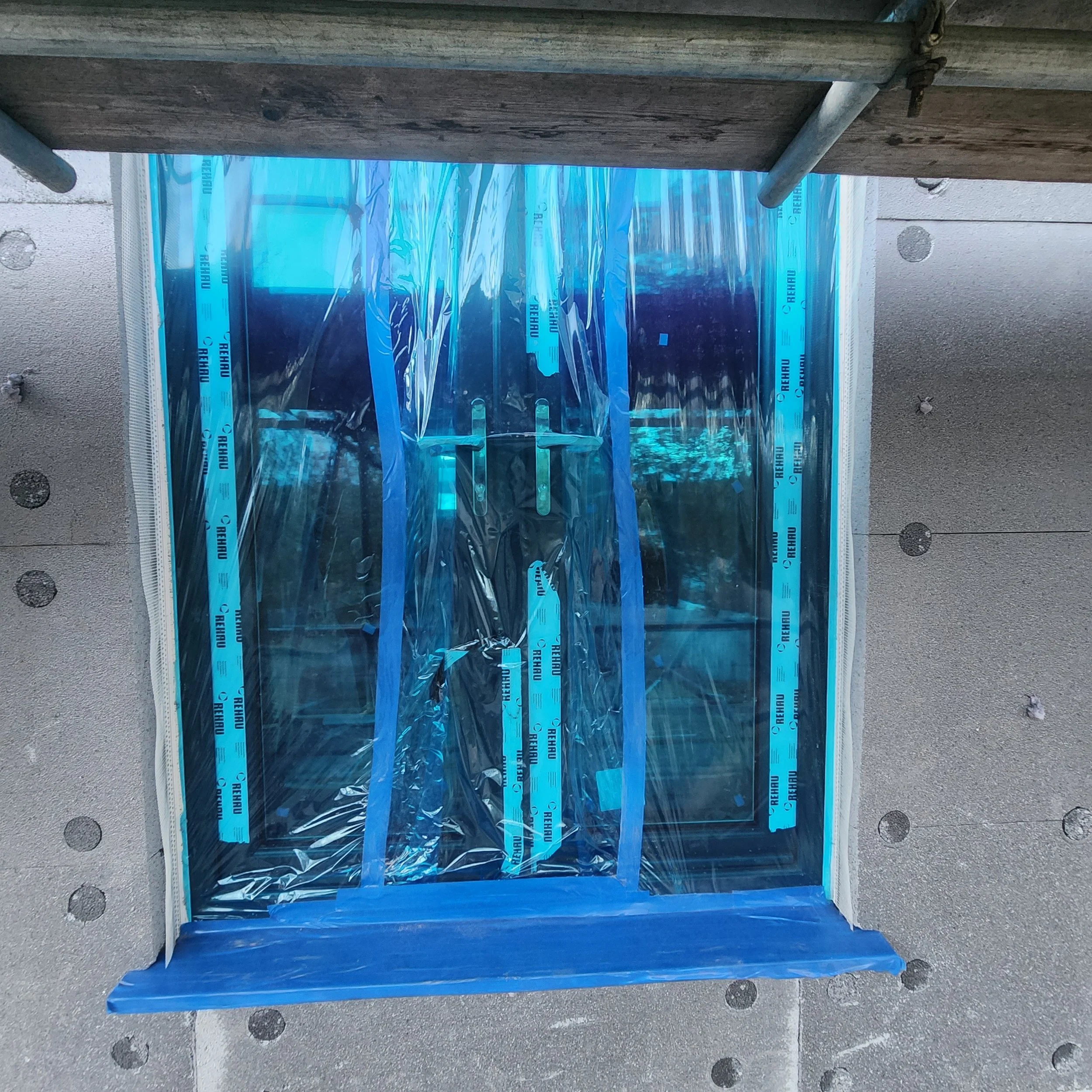 Close-up of a window installation surrounded by beige concrete wall, with blue weather-resistant membrane tape around the edges and plastic sheeting covering the window opening.
