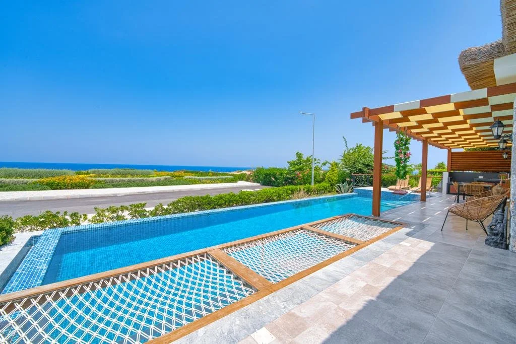 Poolside view with a swimming pool, lounge chairs, and a shaded patio area overlooking the ocean.