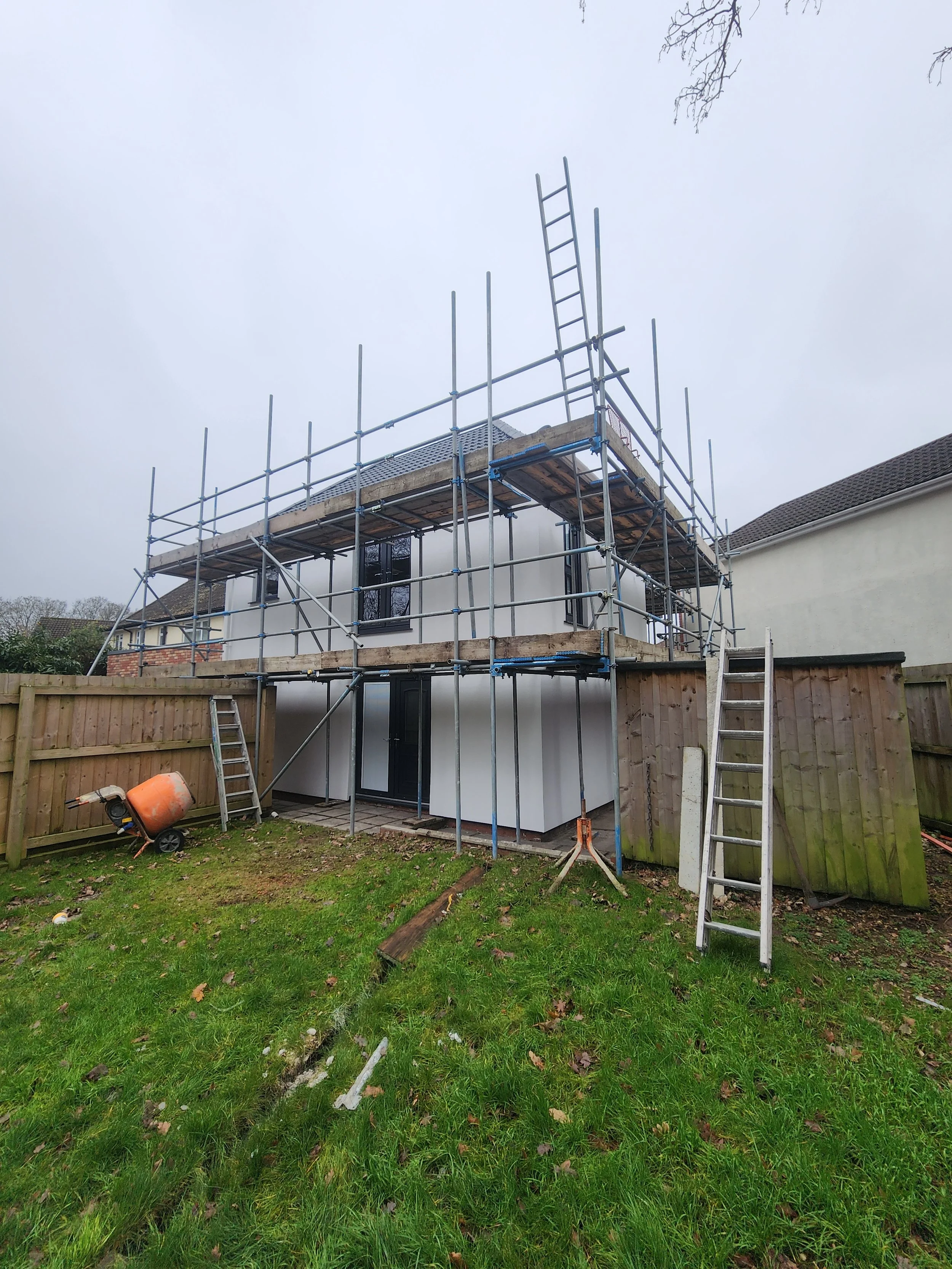 Construction scaffolding around a two-story house with white exterior walls, black doors and windows, on a cloudy day. Tools and ladders are on the ground, and a fence surrounds the yard.