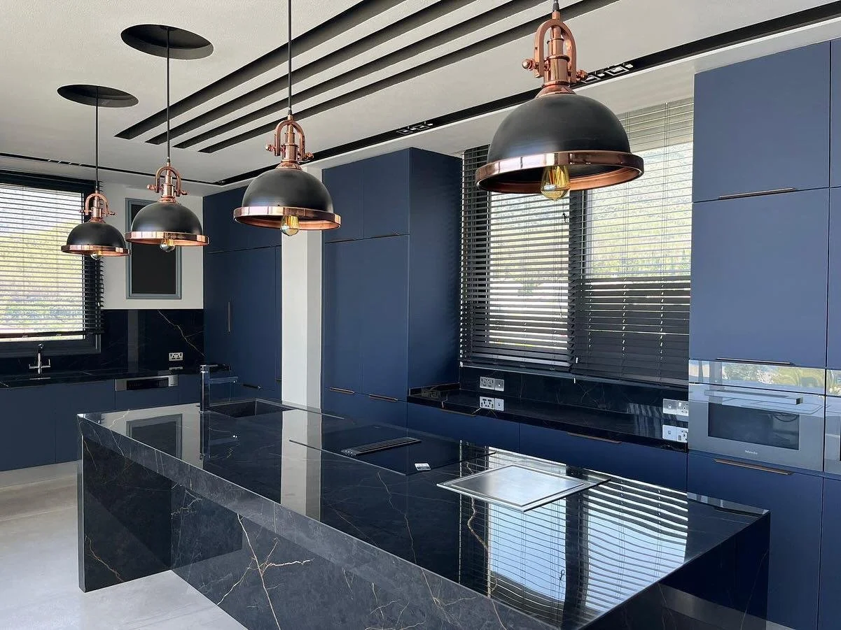 Modern kitchen with blue cabinets, black marble countertops, three black and copper pendant lights, and large windows with blinds.