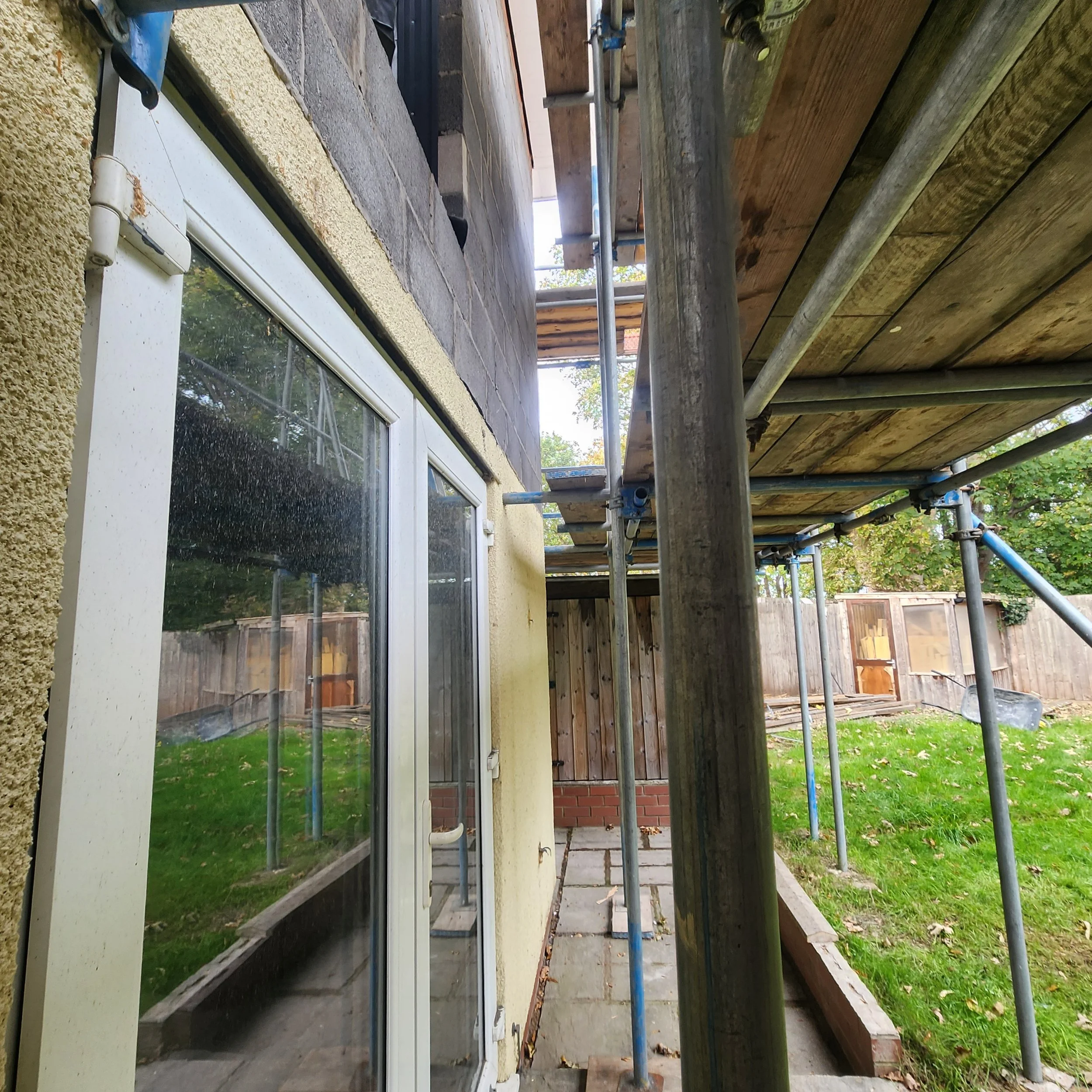 Scaffolding set up around the exterior of a house with a sliding glass door, a paved pathway, a wooden fence, and green grass with fallen leaves.