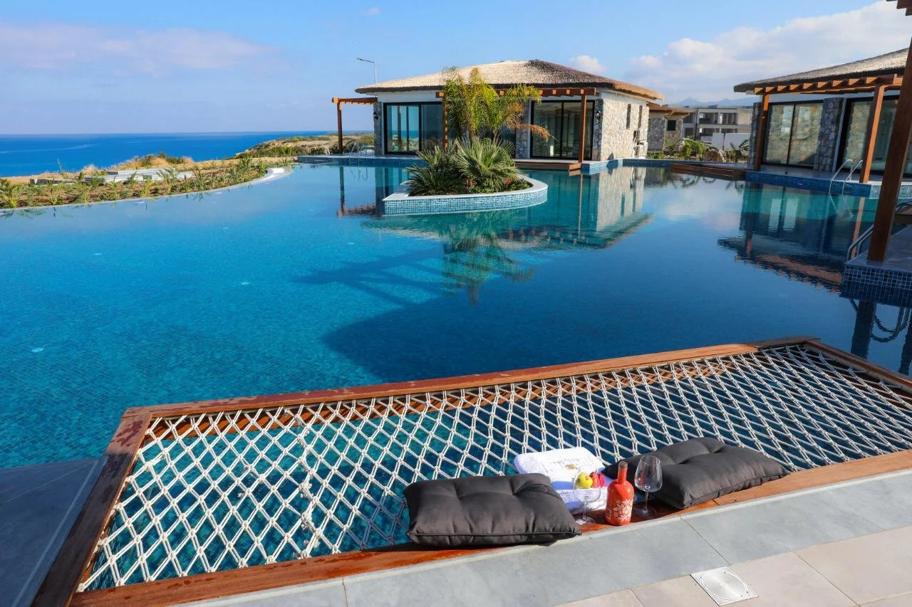 Luxurious infinity pool overlooking the ocean with a wooden lounge chair, black cushions, wine glasses, a bottle, and towels with a tropical resort in the background.