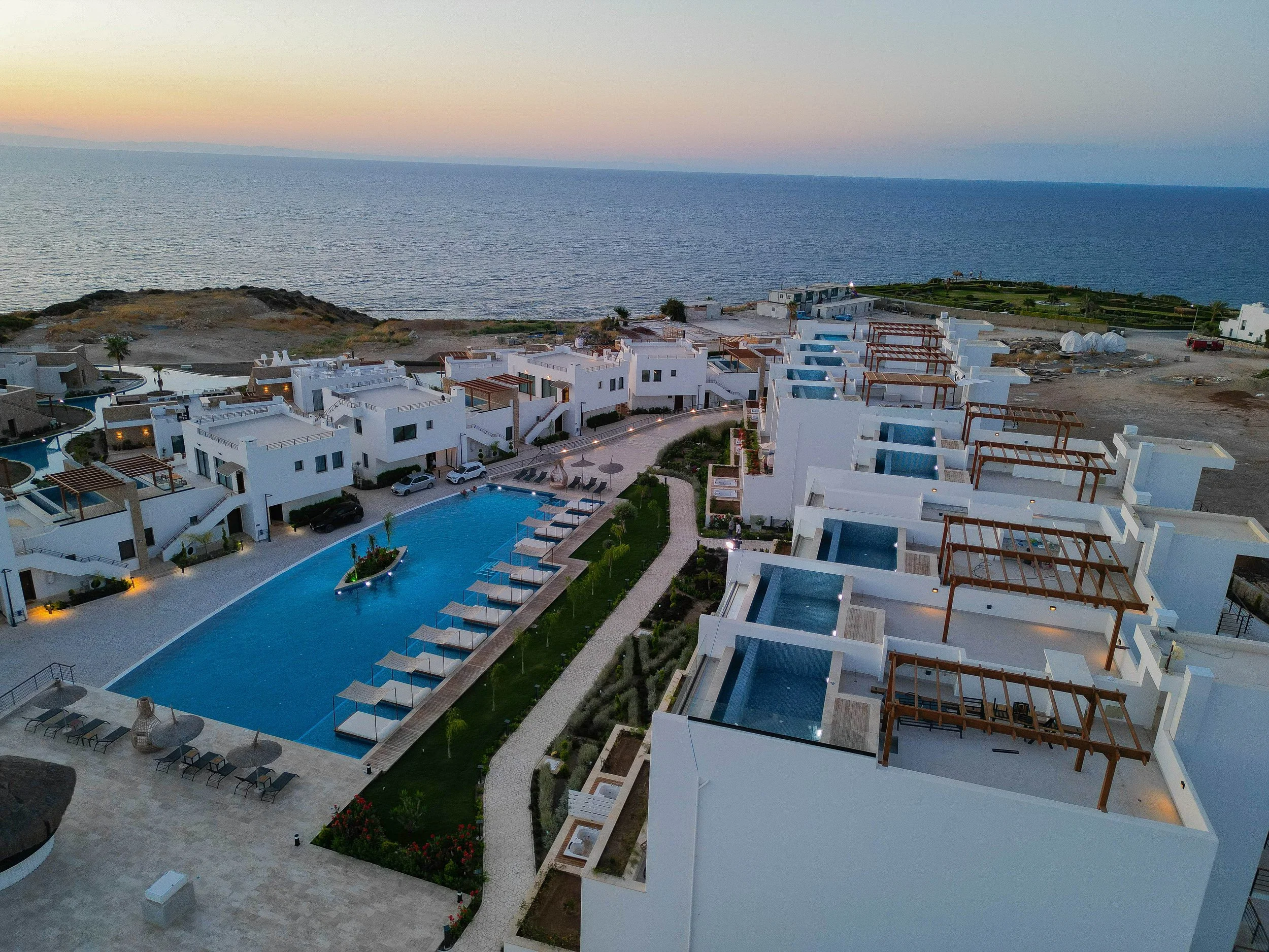 Aerial view of a modern seaside resort with white buildings, swimming pool, lounge chairs, and ocean in the background at sunset.