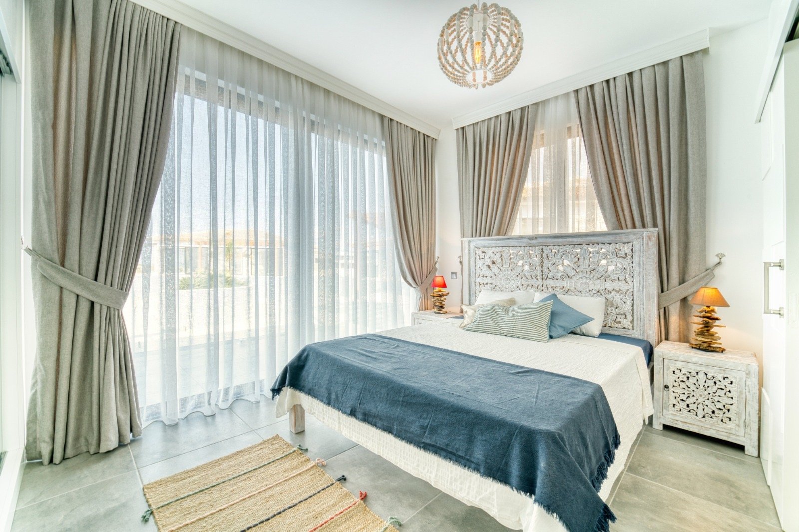 A bright bedroom with large windows covered by sheer curtains and thicker drapes, a white carved headboard, a bed with white and blue bedding, two small bedside tables with lamps, and a woven rug on tiled floor.