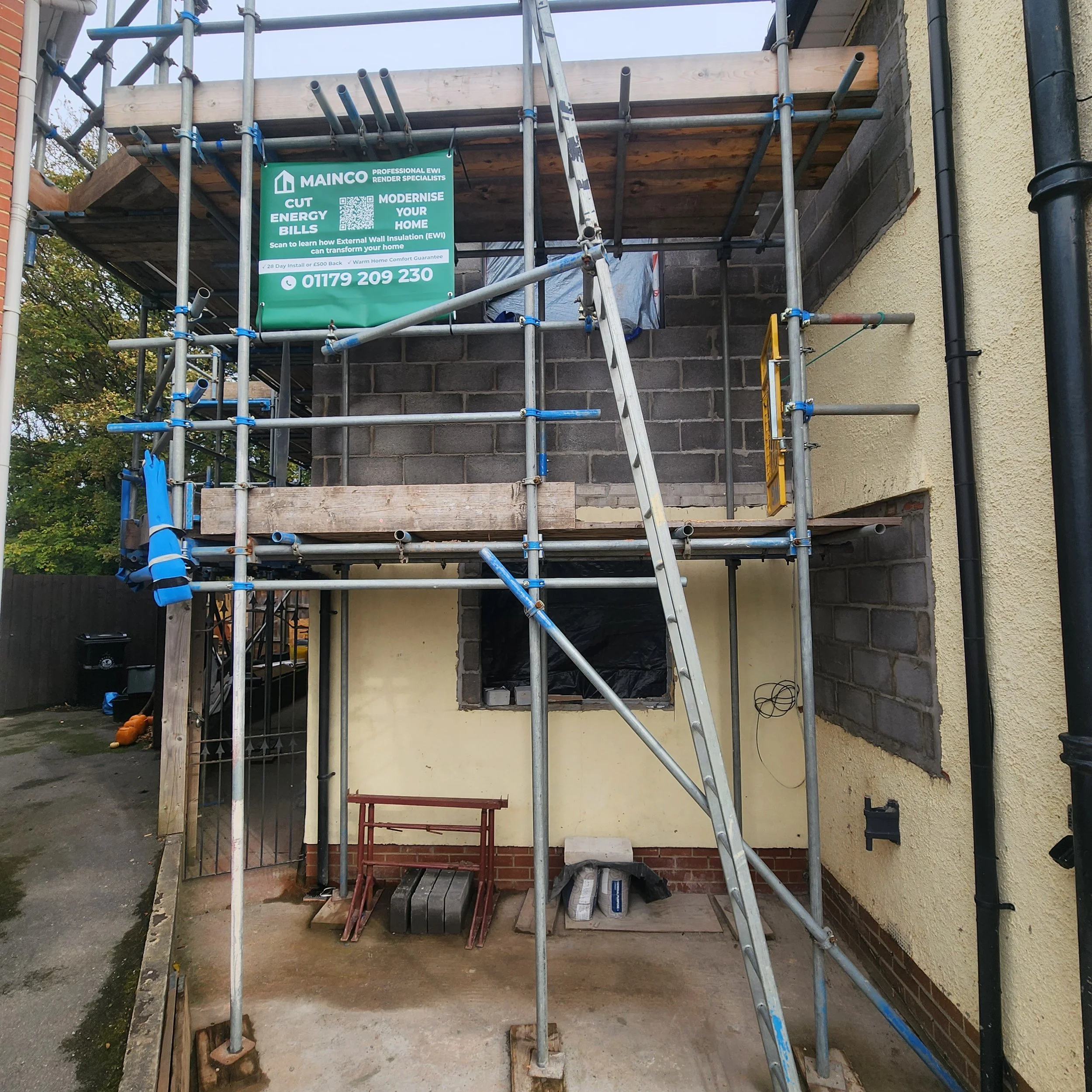 Front of a house under construction with scaffolding in place, showing brickwork and construction materials on the ground.