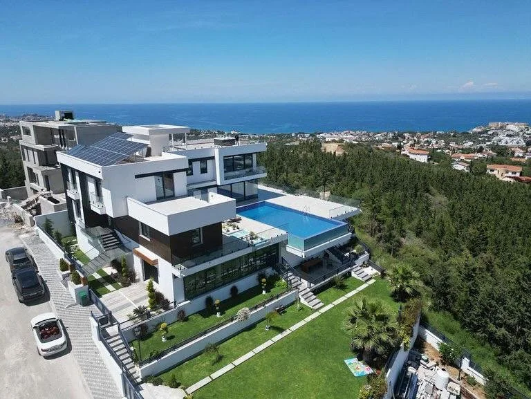Modern multi-story house with swimming pool, surrounded by a well-kept lawn, located on a hillside with ocean view in the background.