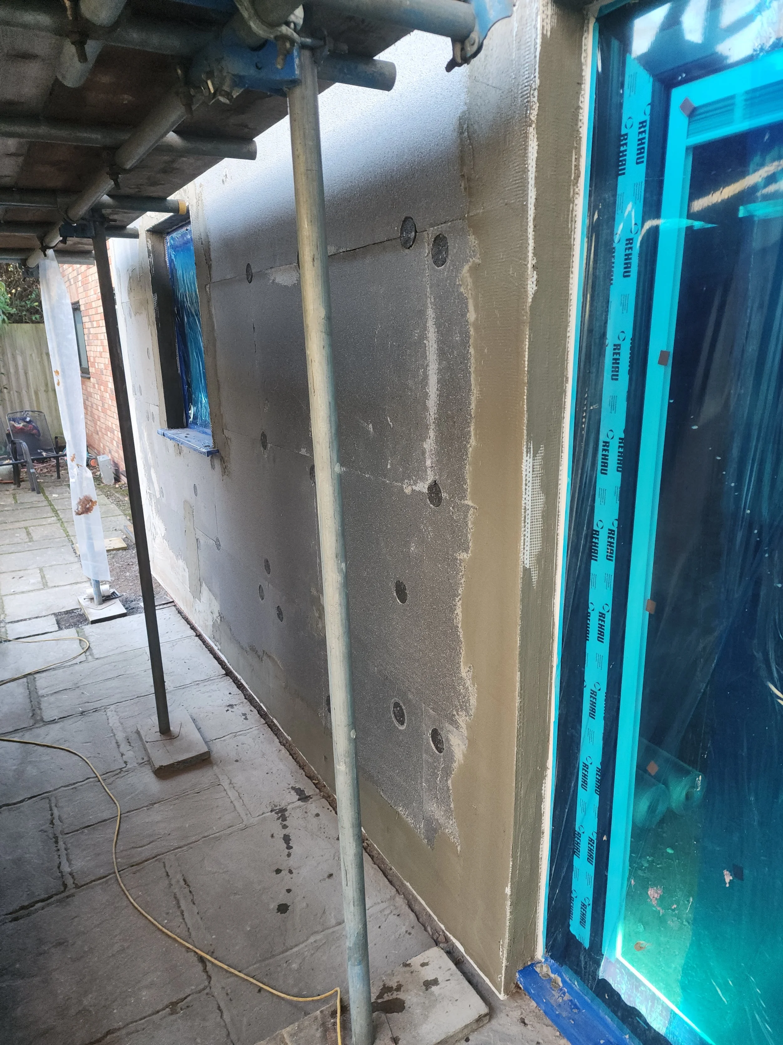 Exterior wall under construction with scaffolding, partially plastered, next to a blue window frame with protective film, and a paved patio.