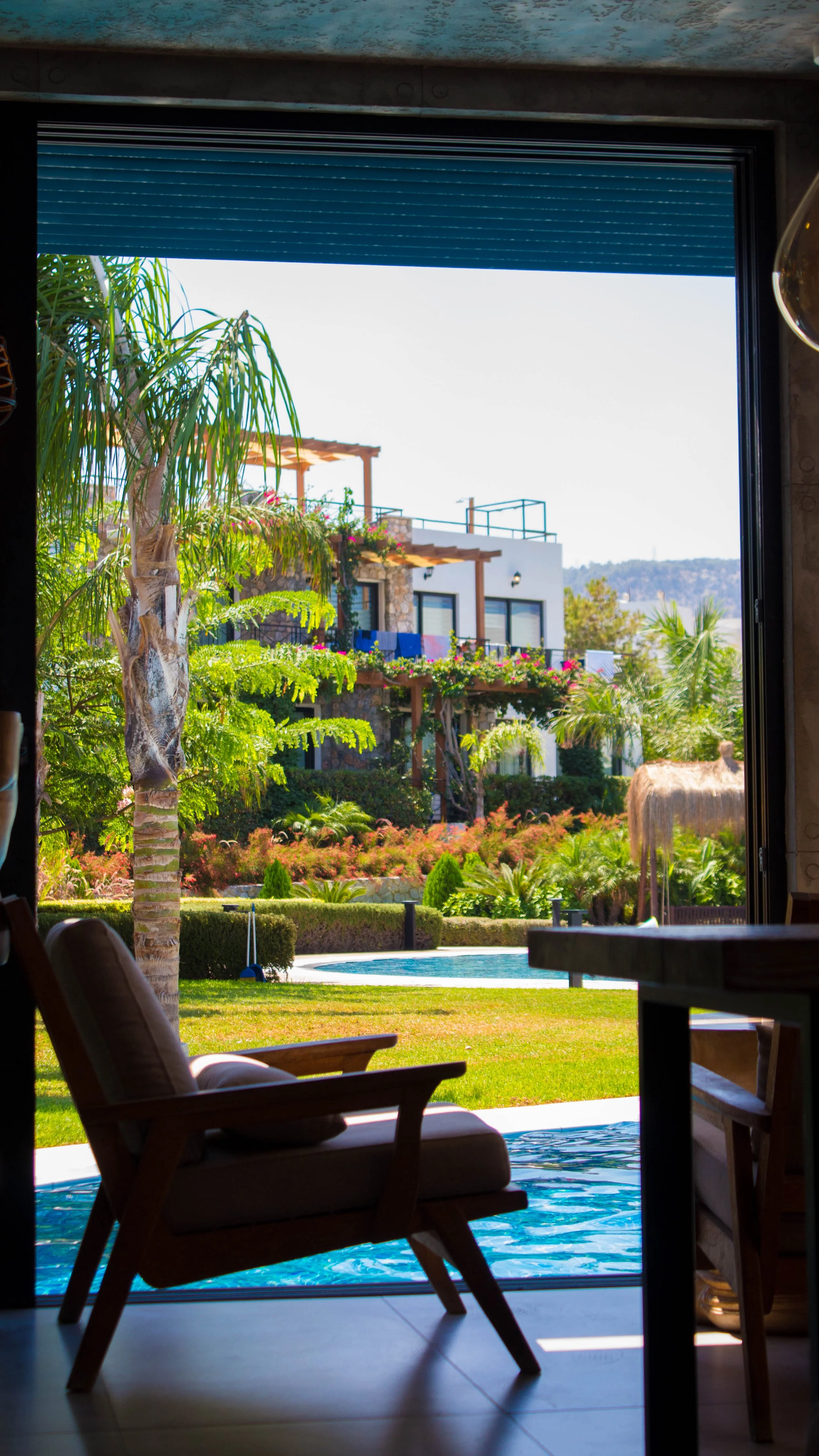 View from inside a house looking out through an open door at a tropical backyard with a swimming pool, lush greenery, palm trees, colorful flowering plants, multi-story white building with balconies, and hills in the background.