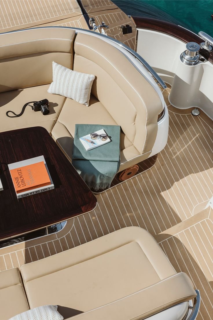 View of a beige leather seating area on a yacht with a black camera, sunglasses, a book, and a magazine on top, and a small pillow on the corner. There is a wooden table and a white cylindrical object with a polished metal handle and a metal base, ne