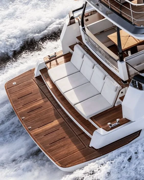 Aerial view of a yacht with a white cushioned seating area on the deck, surrounded by wooden flooring, moving through the water.