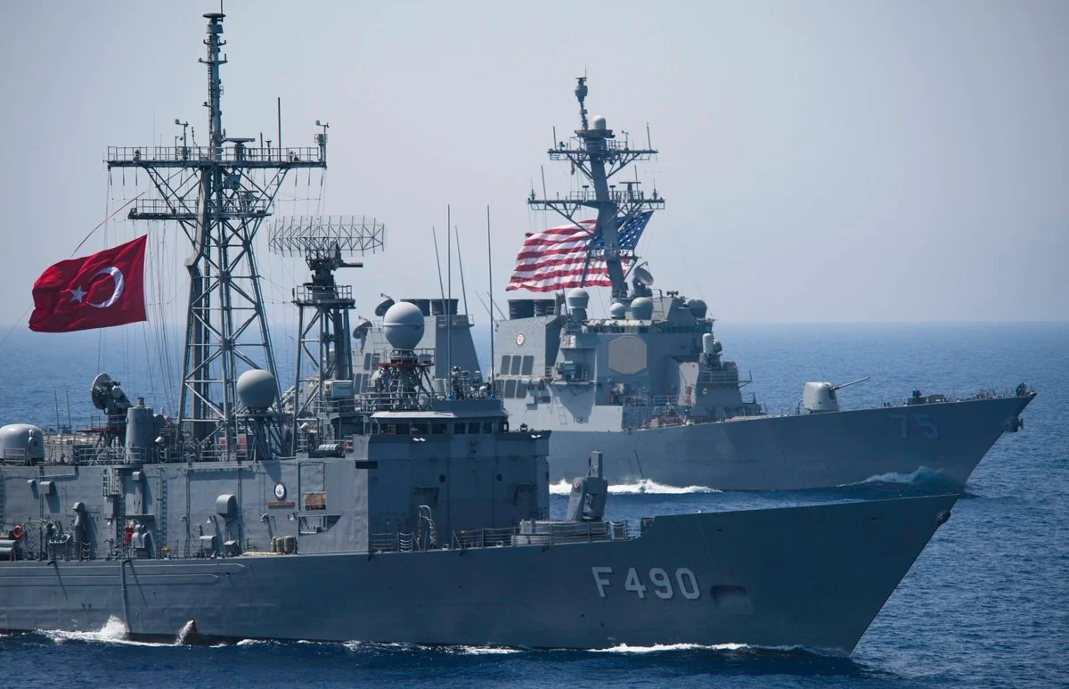 Two military ships sailing in the ocean, with flags flying; one ship in the foreground with the number F490, and a larger ship with the number 75 and an American flag in the background.