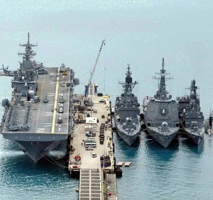 Four large naval ships docked at a pier with vehicles and equipment on the dock, and a crane visible.